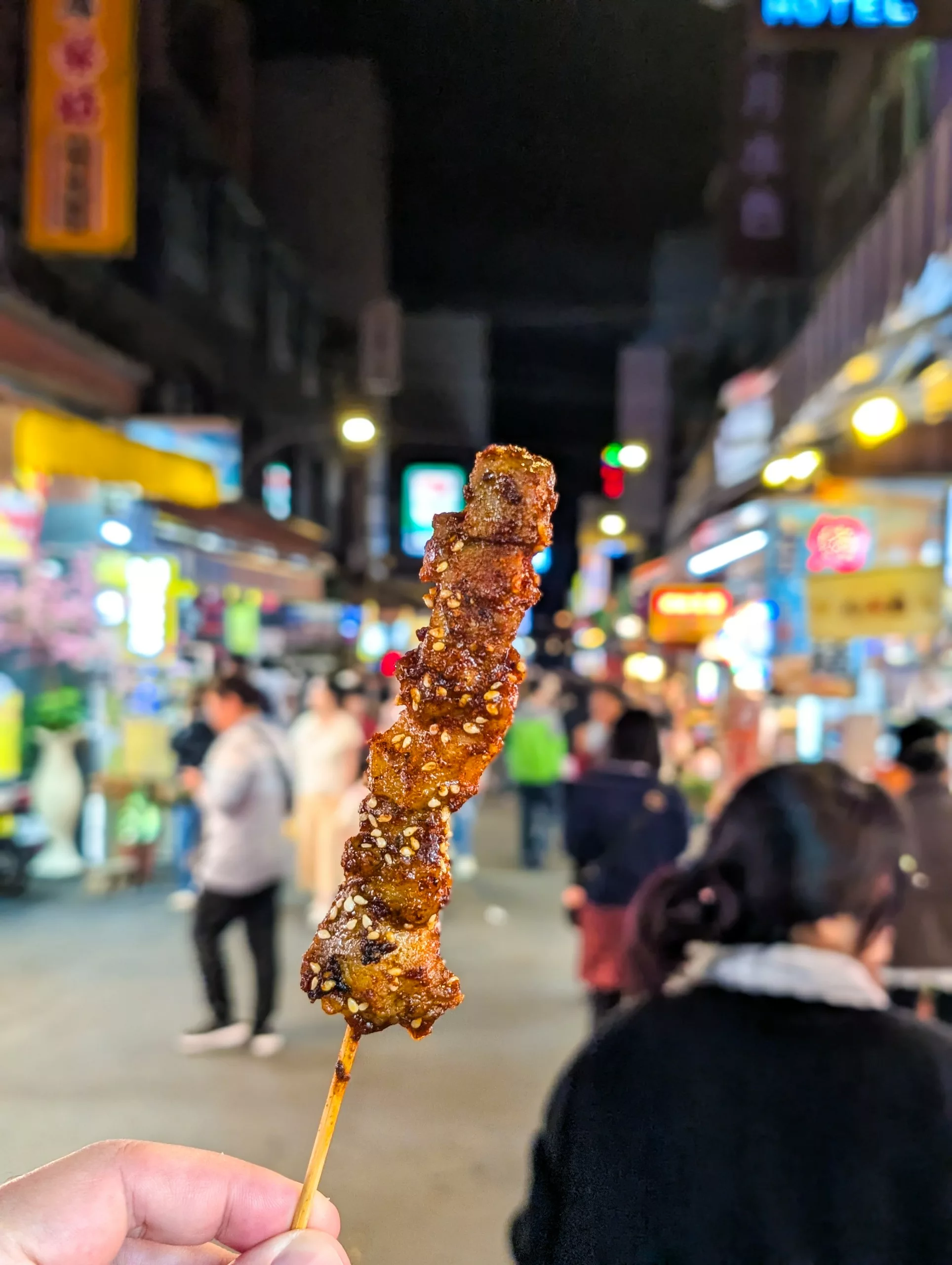 Une main tient une brochette de viande grillée en pleine nuit, avec en arrière-plan un marché de rue animé et très éclairé et des personnes floues