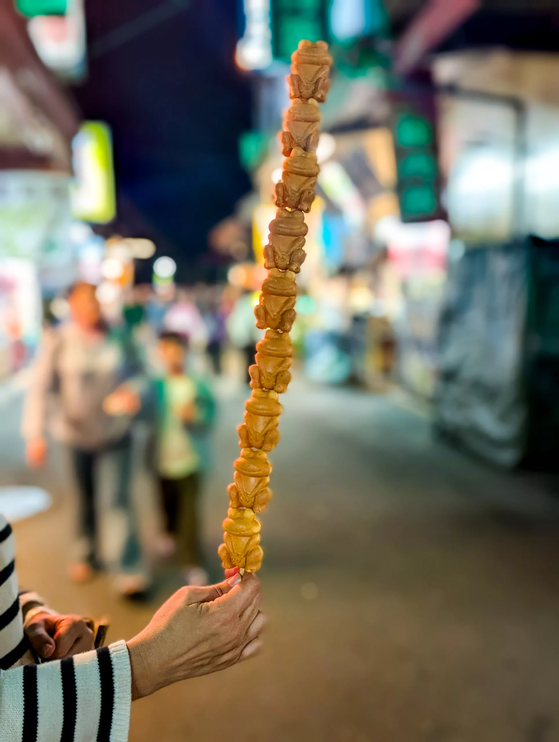 Un visiteur tient une brochette de boulettes en forme de grenouilles frites brun doré, au milieu de personnes floues et de lumières scintillantes en arrière-plan.