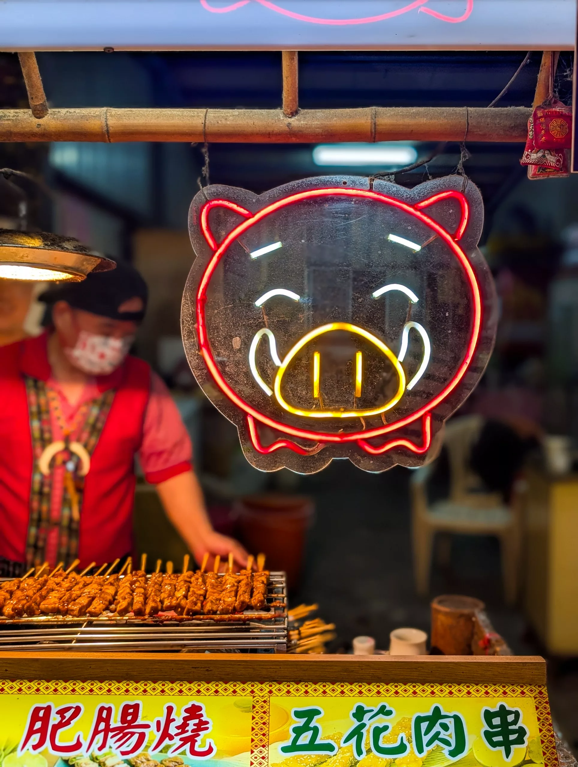 Un stand de nourriture présente des brochettes grillées devant une enseigne en néon représentant un cochon. Une personne vêtue d'une chemise rouge, d'un tablier et d'un masque se tient derrière les brochettes, avec des signes en chinois en bas.