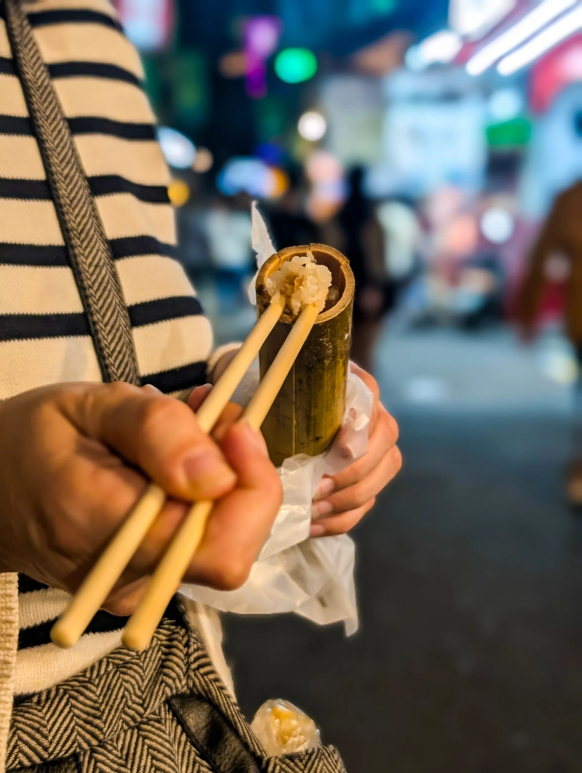 Une personne tient un tube de bambou rempli de riz à l'aide de baguettes, dans un décor extérieur flou et animé.