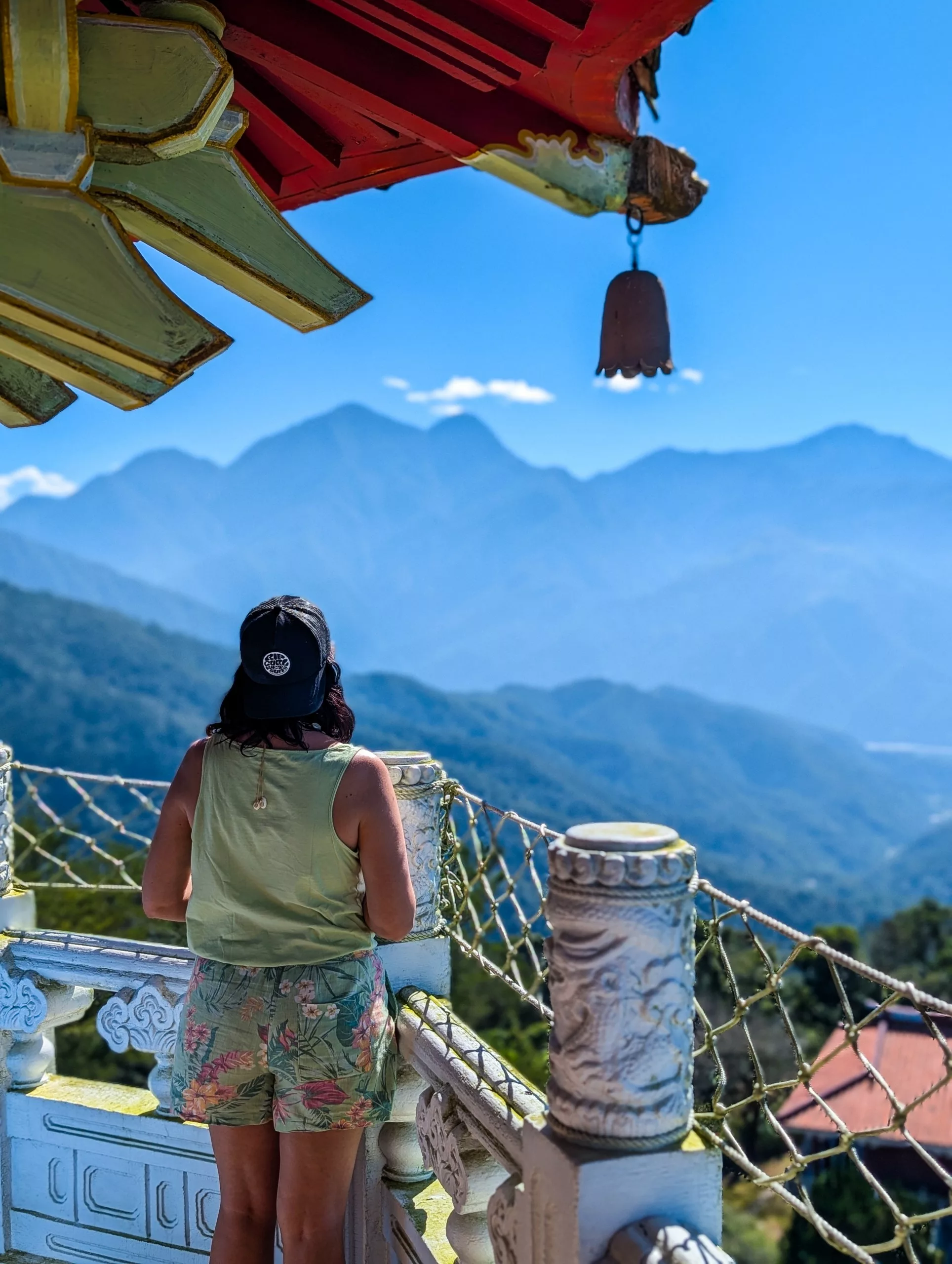 Un visiteur vêtu d'un débardeur, d'un short et d'une casquette se tient sur un balcon orné de sculptures et contemple une chaîne de montagnes. Une cloche suspendue et un toit décoratif sont visibles au-dessus.