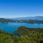 Un grand lac bleu - le Sun Moon Lake - entouré de collines vertes et boisées, avec des montagnes lointaines et quelques bâtiments visibles le long de la rive.