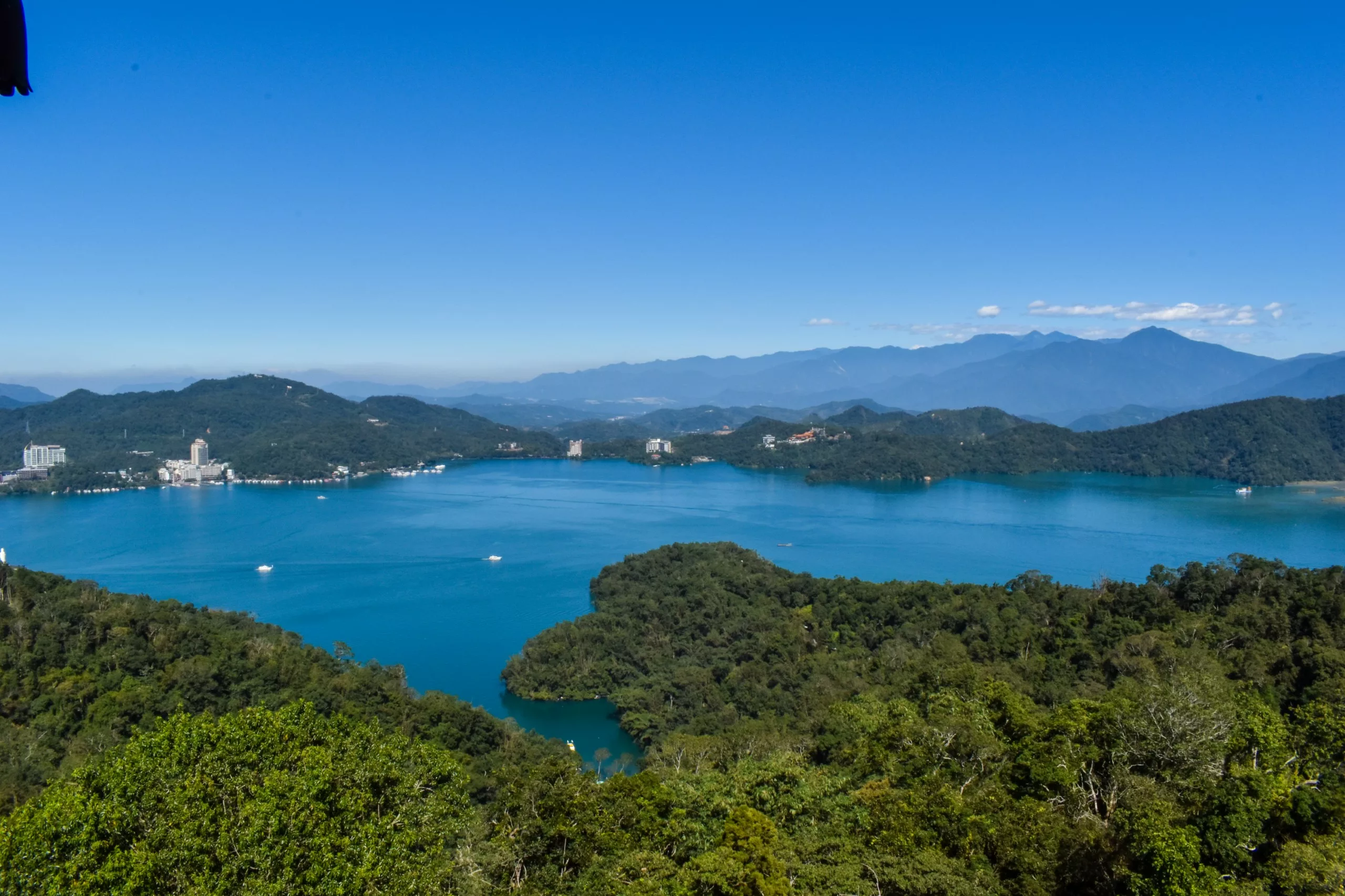 Un grand lac bleu - le Sun Moon Lake - entouré de collines vertes et boisées, avec des montagnes lointaines et quelques bâtiments visibles le long de la rive.