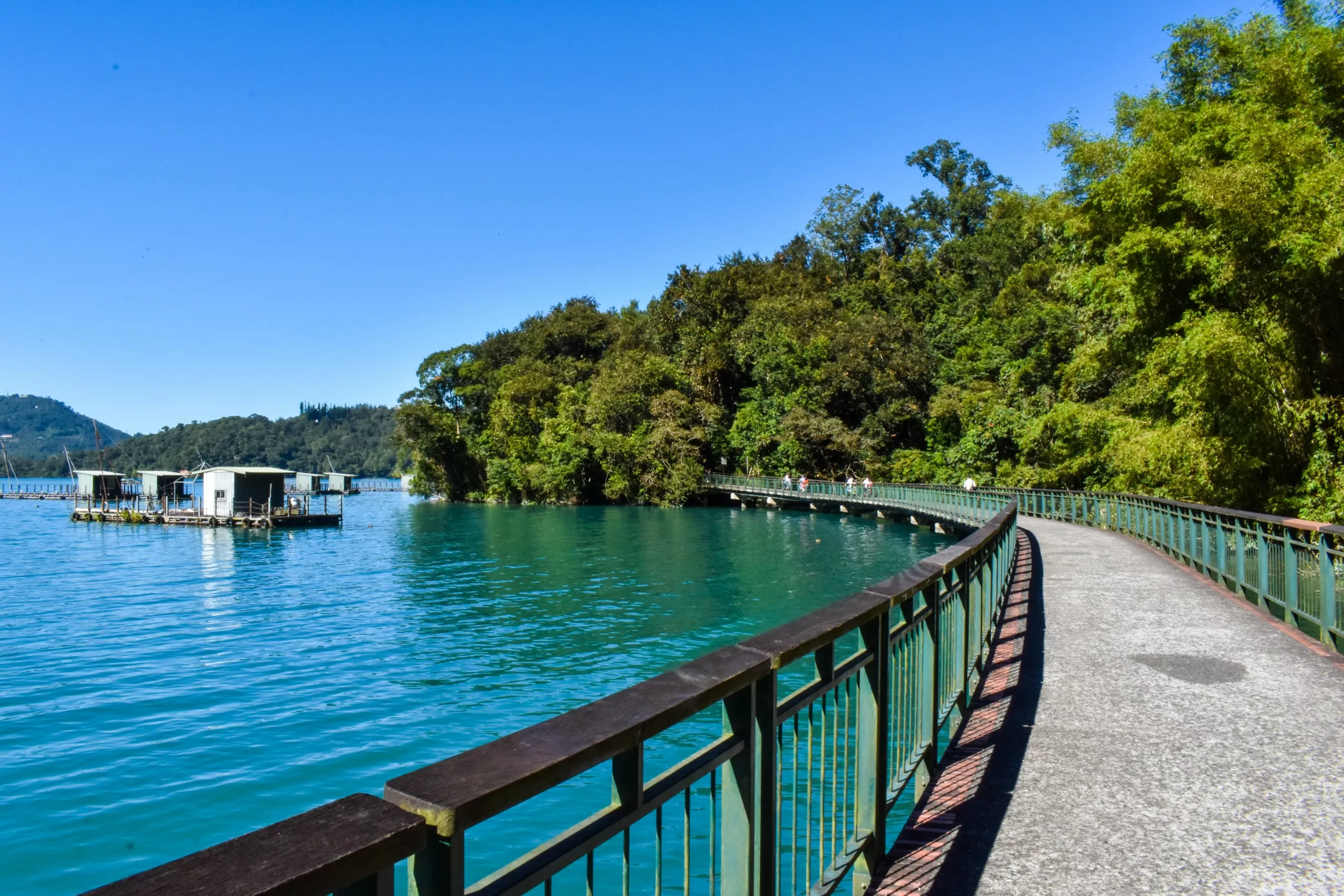 Une allée pavée avec une balustrade verte s'enroule le long du lac Sun Moon, bordée d'arbres verts et denses. Des structures flottantes sont ancrées sur l'eau sous un ciel bleu clair.