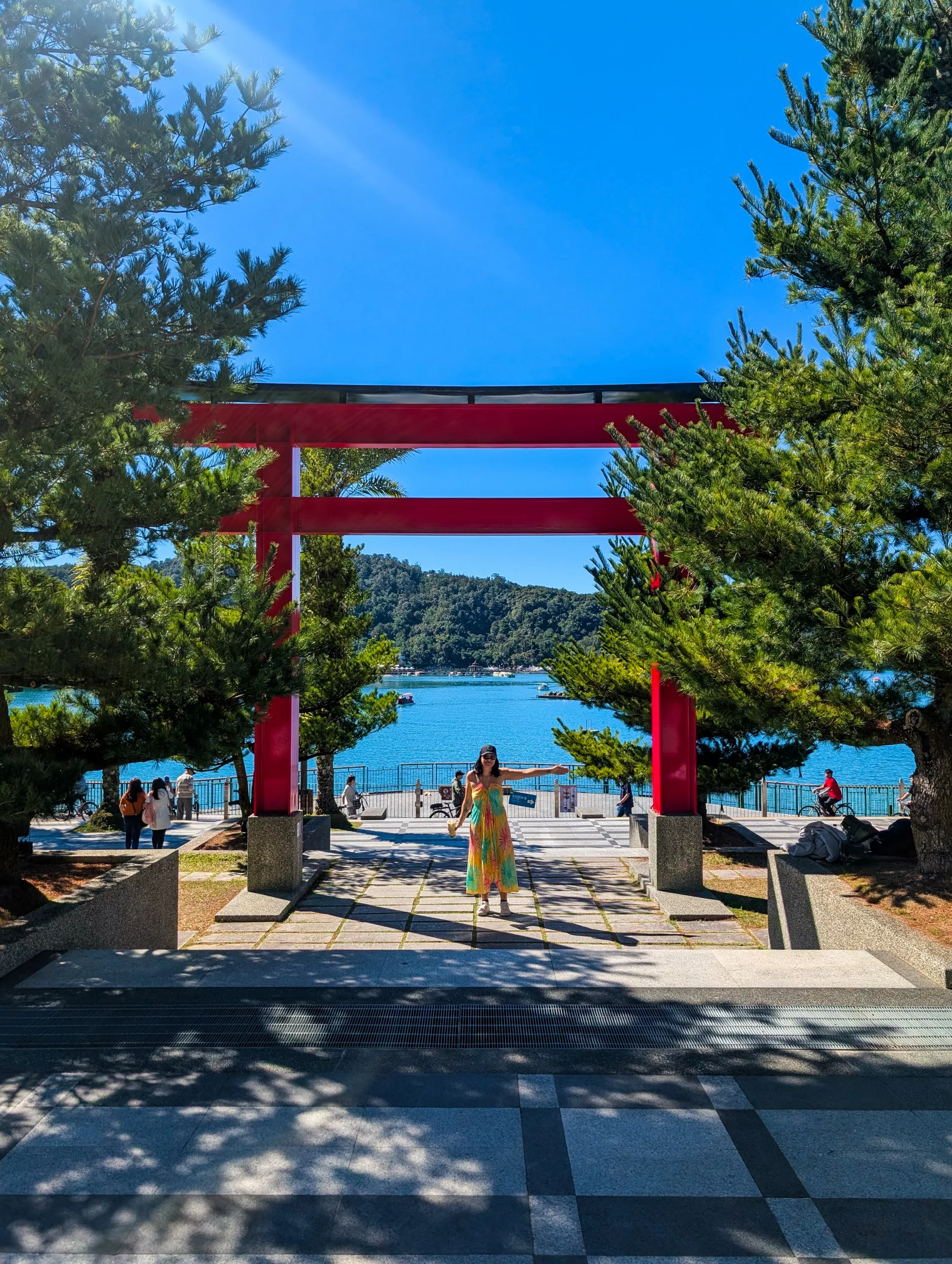 Une visiteuse vêtue d'une robe colorée se tient sous une grande porte torii rouge, près de l'eau du lac Sun Moon, entourée de pins et d'autres arbres, avec un ciel bleu et des collines boisées en arrière-plan.