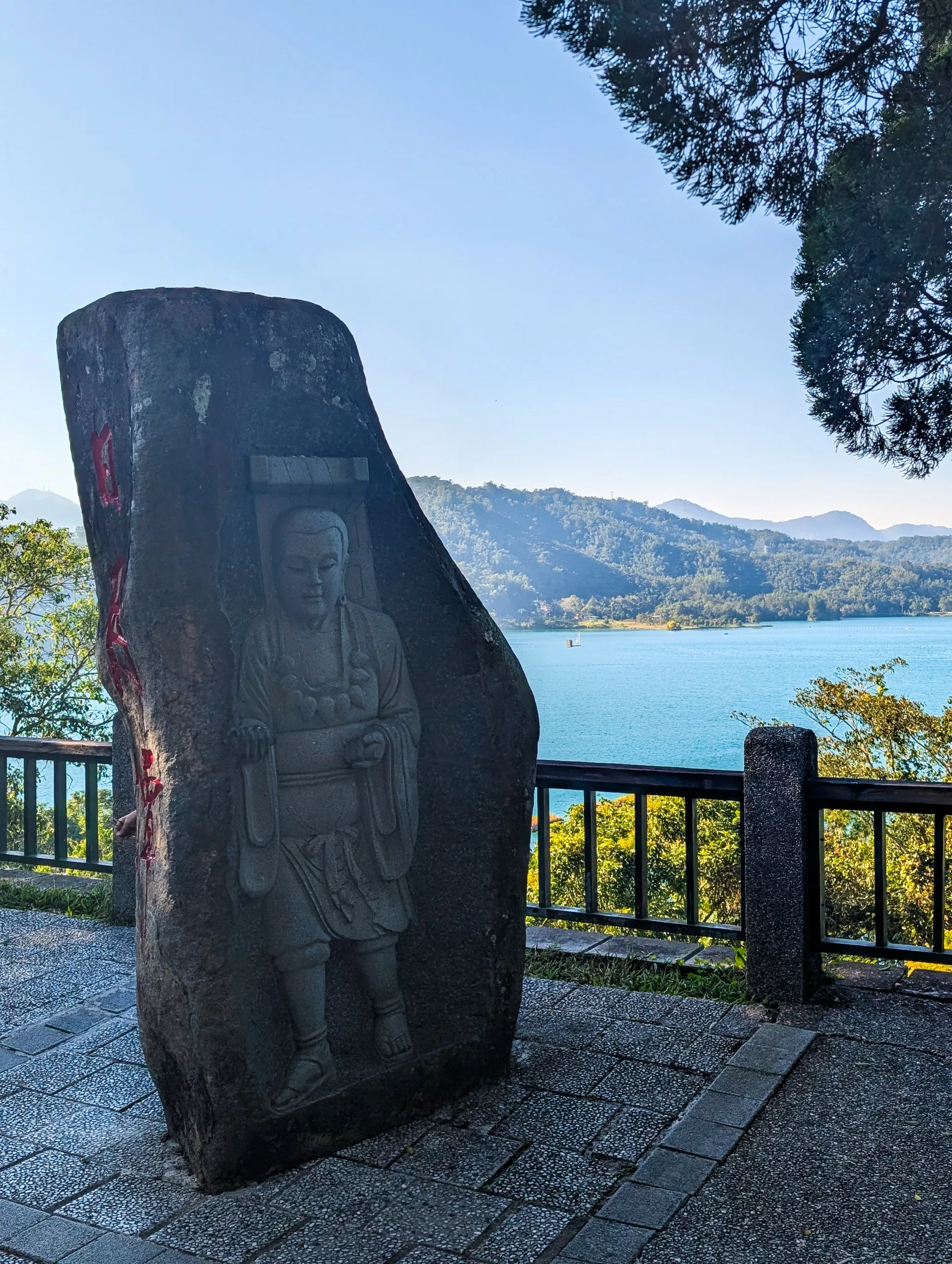 Une sculpture en pierre représentant un personnage debout est encastrée dans un gros rocher près d'une balustrade en métal, surplombant le lac Sun Moon Lake avec des collines boisées et des montagnes au loin sous un ciel dégagé