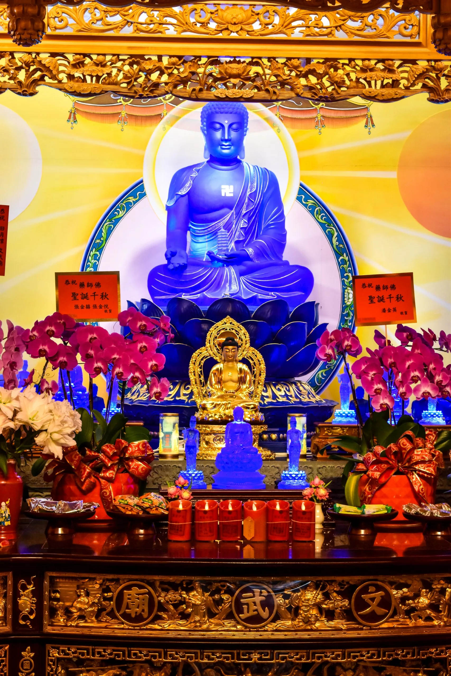 Un vibrant autel bouddhiste avec une grande statue de Bouddha bleue au centre, entouré de statues dorées, d'orchidées violettes, d'inscriptions rouges, de décorations ornementales et d'offrandes sur une table dorée et noire.