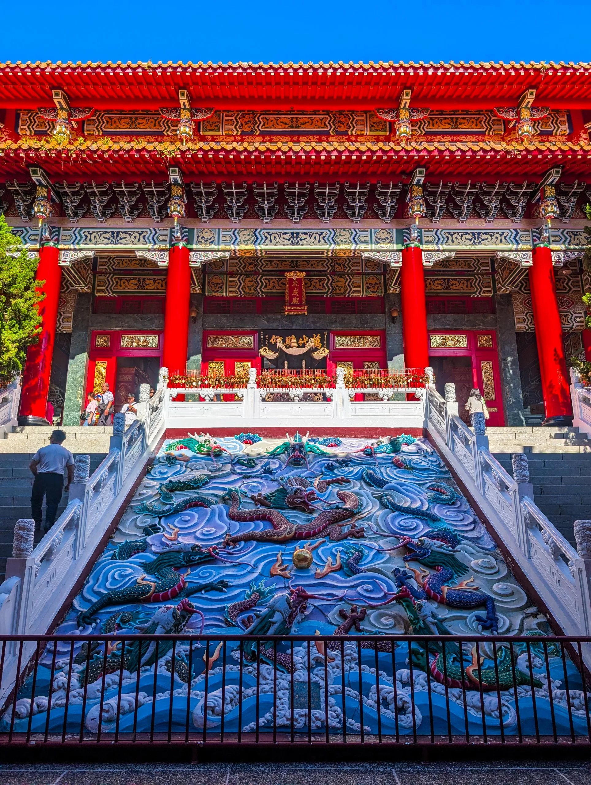 Un temple chinois traditionnel présente des colonnes rouges, des détails de toiture ornés et un large escalier orné d'un dragon en relief coloré. Plusieurs visiteurs se tiennent debout ou marchent près des marches lors d'une journée ensoleillée.