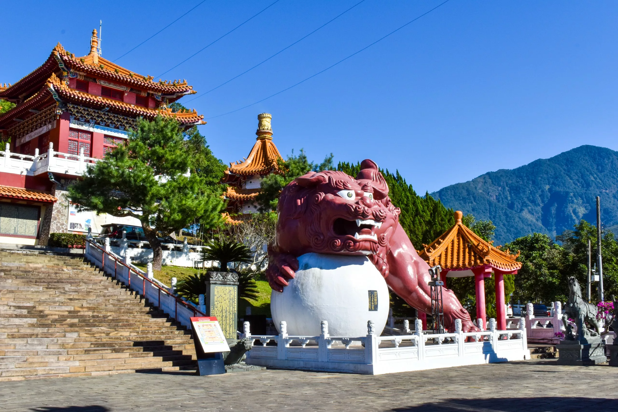Une grande statue de chien foo rouge avec une patte sur une sphère blanche se trouve à côté de marches en pierre menant à un temple chinois traditionnel