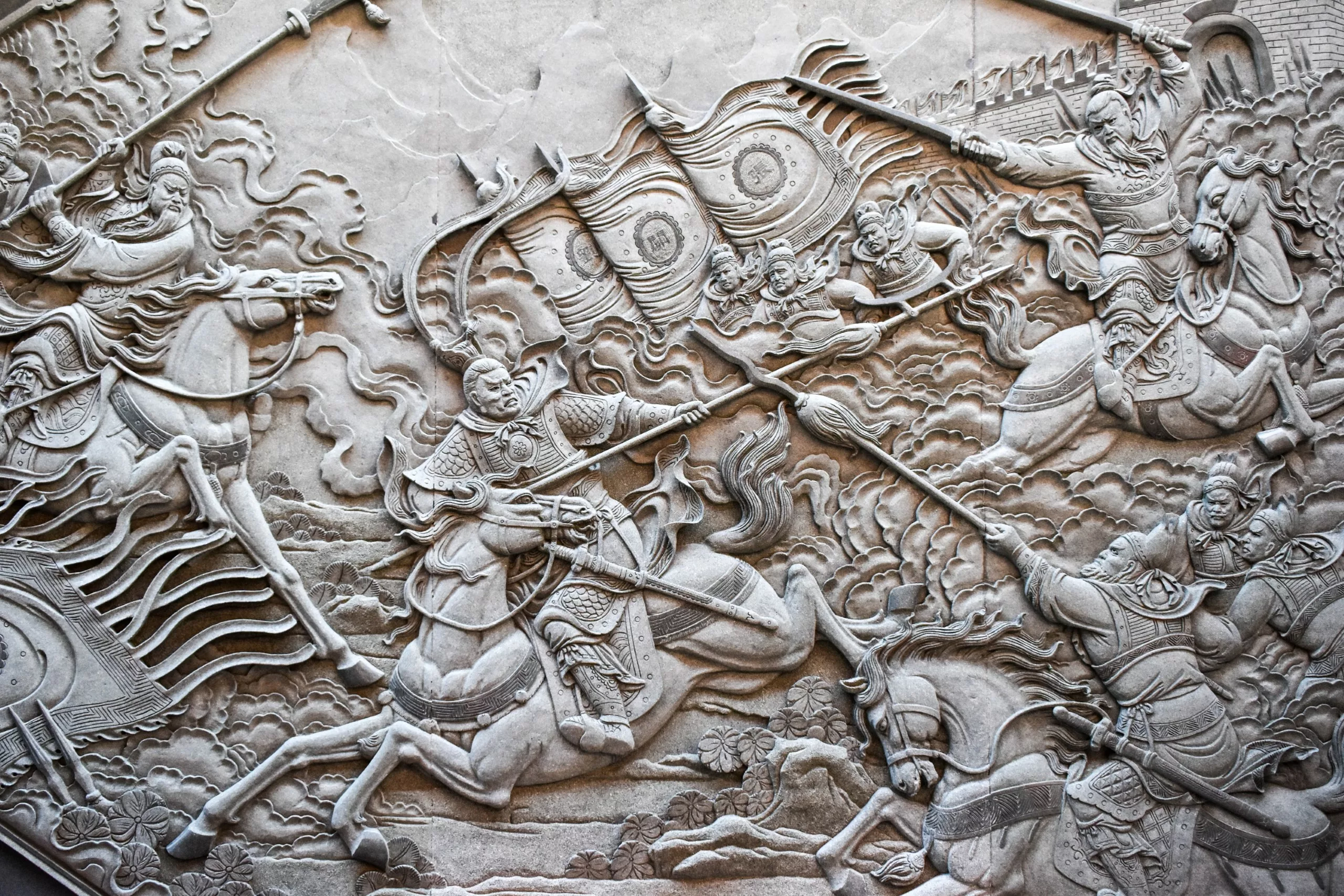 Sculpture en relief en pierre représentant une scène de bataille intense avec des guerriers à cheval, des bannières tourbillonnantes et des flammes sur le mur d'une forteresse