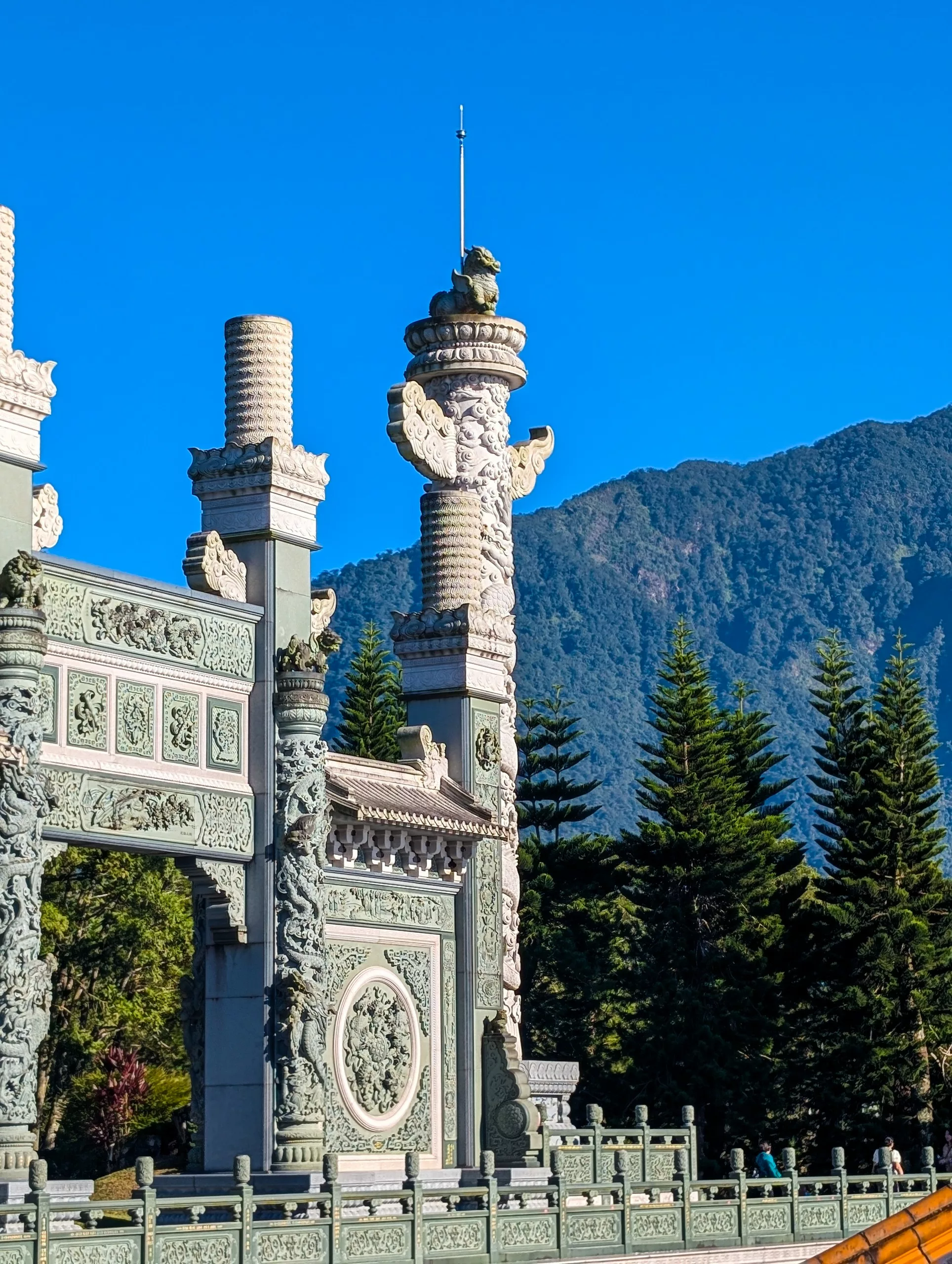 Une arche de pierre ornée de sculptures et de colonnes complexes se dresse devant des pins, avec un ciel bleu clair et des montagnes boisées