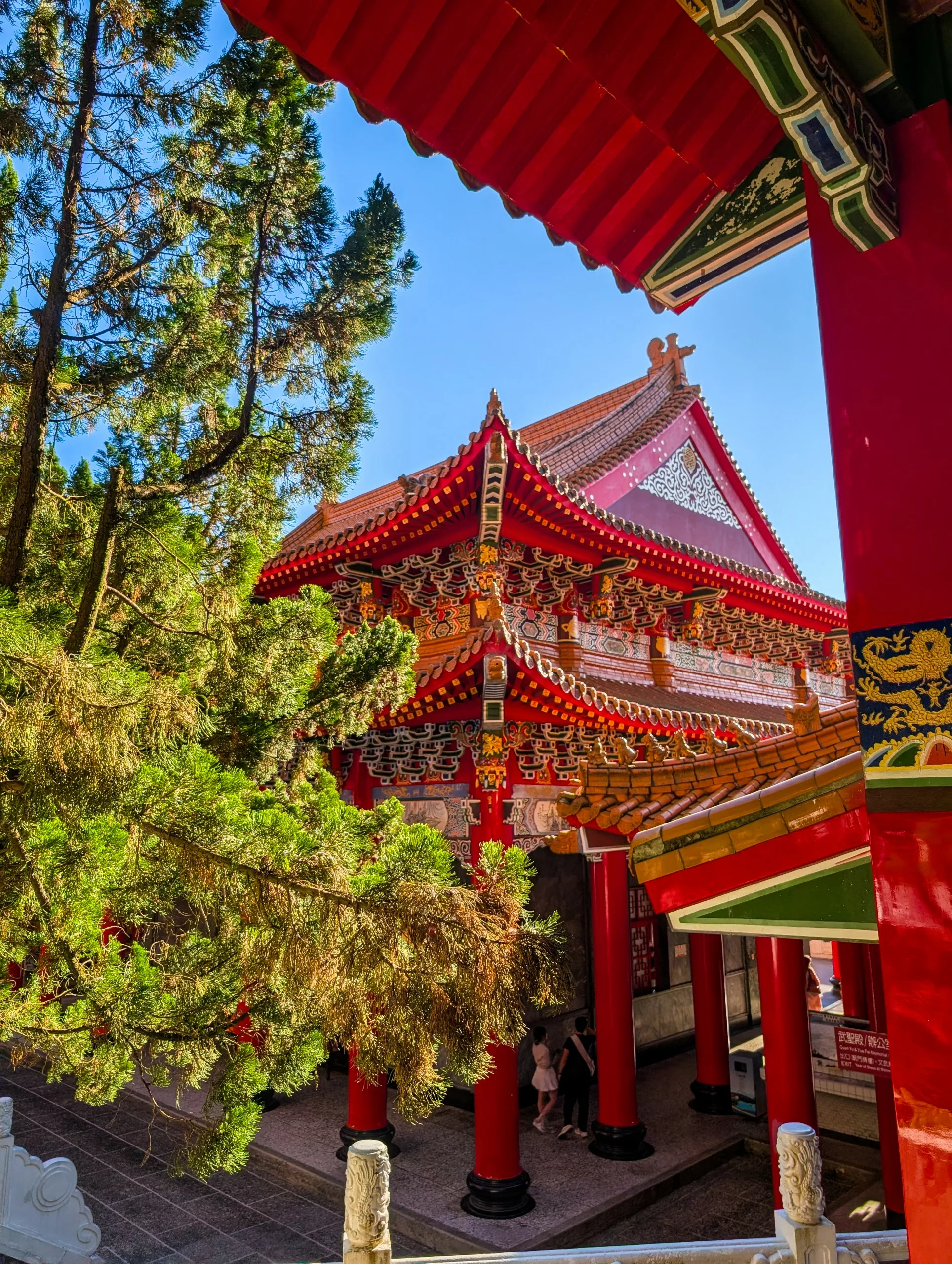 Un temple chinois traditionnel avec des piliers rouges ornés et un toit à plusieurs niveaux avec des détails complexes en or et en rouge, entouré de grands arbres verts sous un ciel bleu clair.