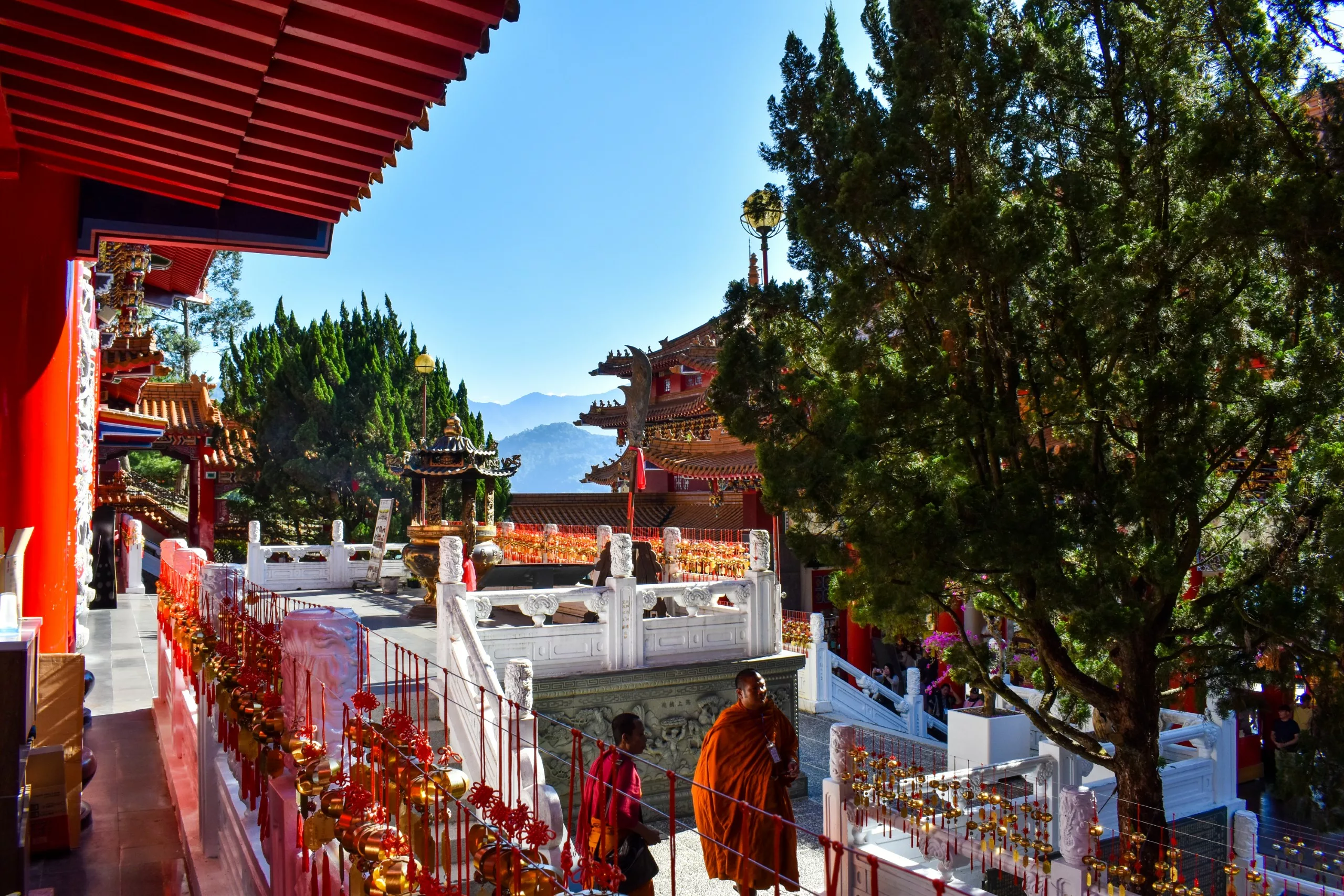 Un moine bouddhiste en robe orange marche près de la cour d'un temple traditionnel aux colonnes et sculptures rouges. Des montagnes s'élèvent à l'arrière-plan sous un ciel bleu clair.