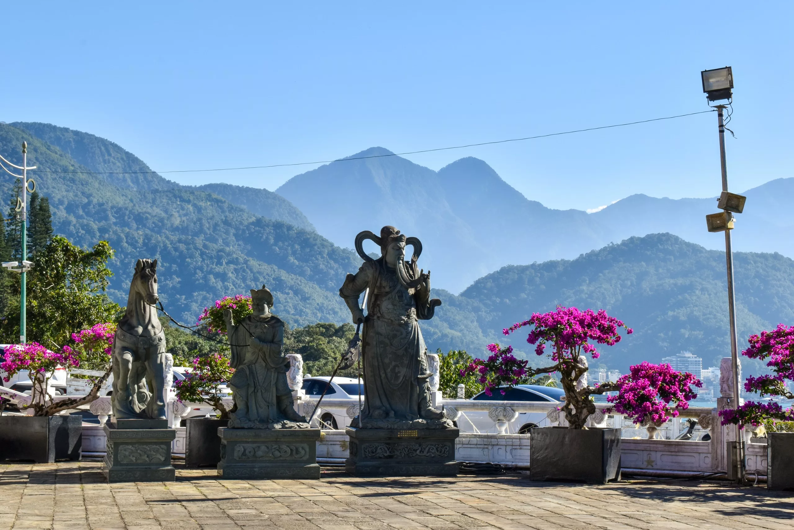 Trois statues de pierre se dressent sur une terrasse carrelée avec des fleurs roses en pot, entourées de montagnes vertes et d'un ciel bleu
