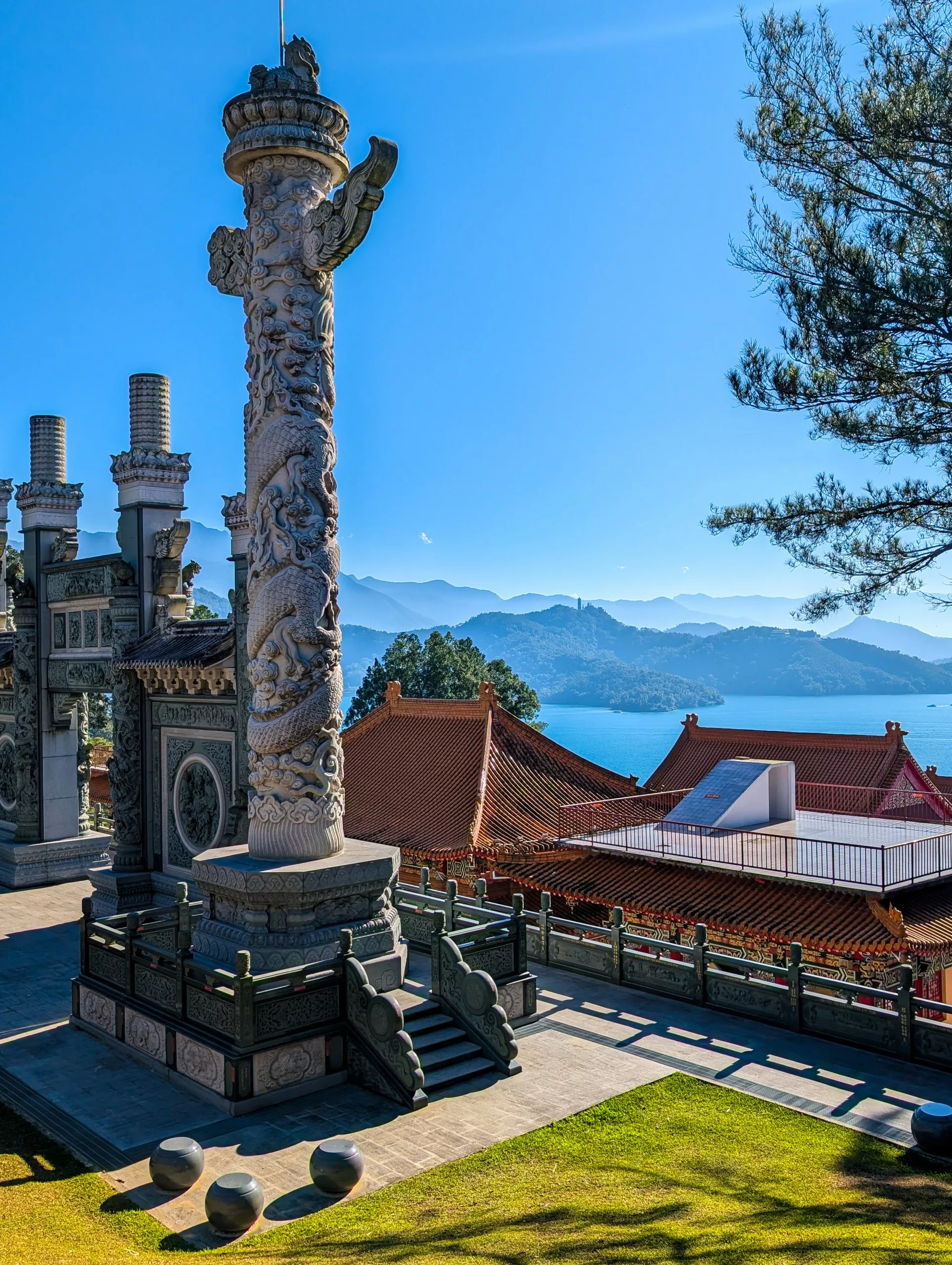 Un haut pilier de pierre sculpté de façon complexe se dresse à côté de temples aux toits de tuiles rouges, surplombant le Sun Moon Lake et des montagnes lointaines sous un ciel clair. Des branches de pin sont visibles