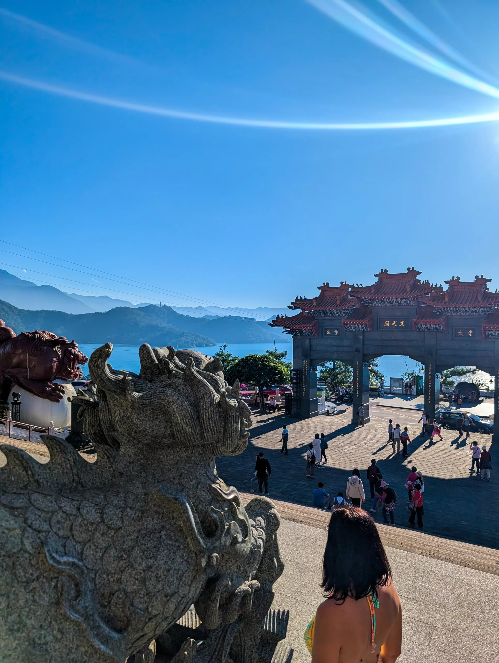 Un visiteur se tient près d'une grande statue de dragon en pierre, surplombant une arche chinoise traditionnelle aux toits rouges au Sun Moon Lake. Des gens se promènent sur la place ouverte avec les montagnes en arrière-plan.