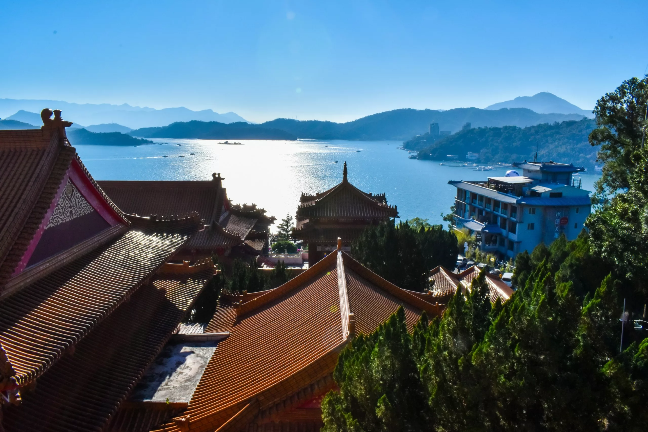 Au premier plan, les toits d'un temple traditionnel chinois surplombent un lac,, avec un bâtiment moderne sur la droite, des arbres et des montagnes lointaines sous un ciel bleu limpide.