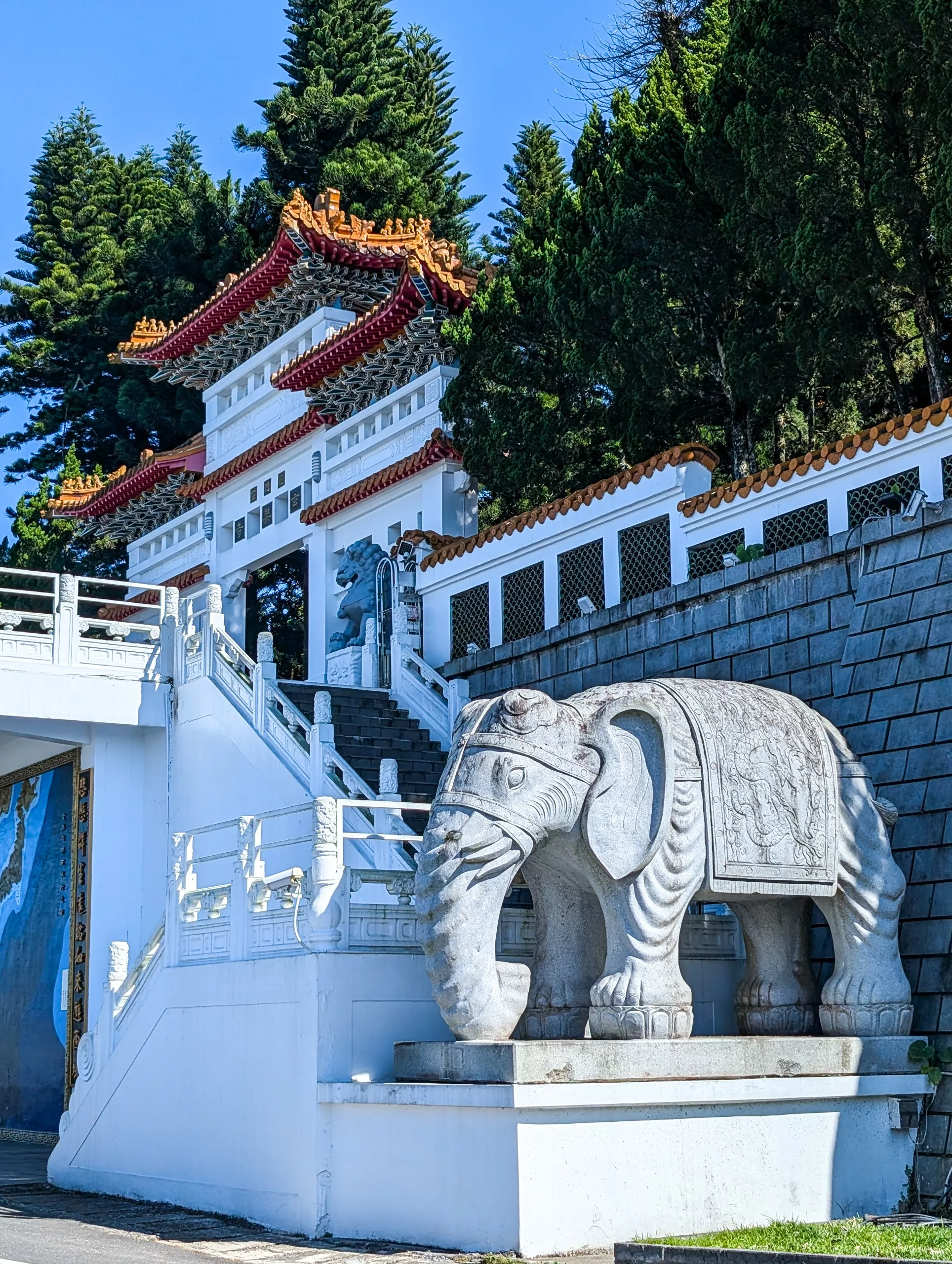 Une grande statue d'éléphant en pierre blanche se trouve à la base d'un escalier menant à une porte traditionnelle avec des arbres verts derrière.