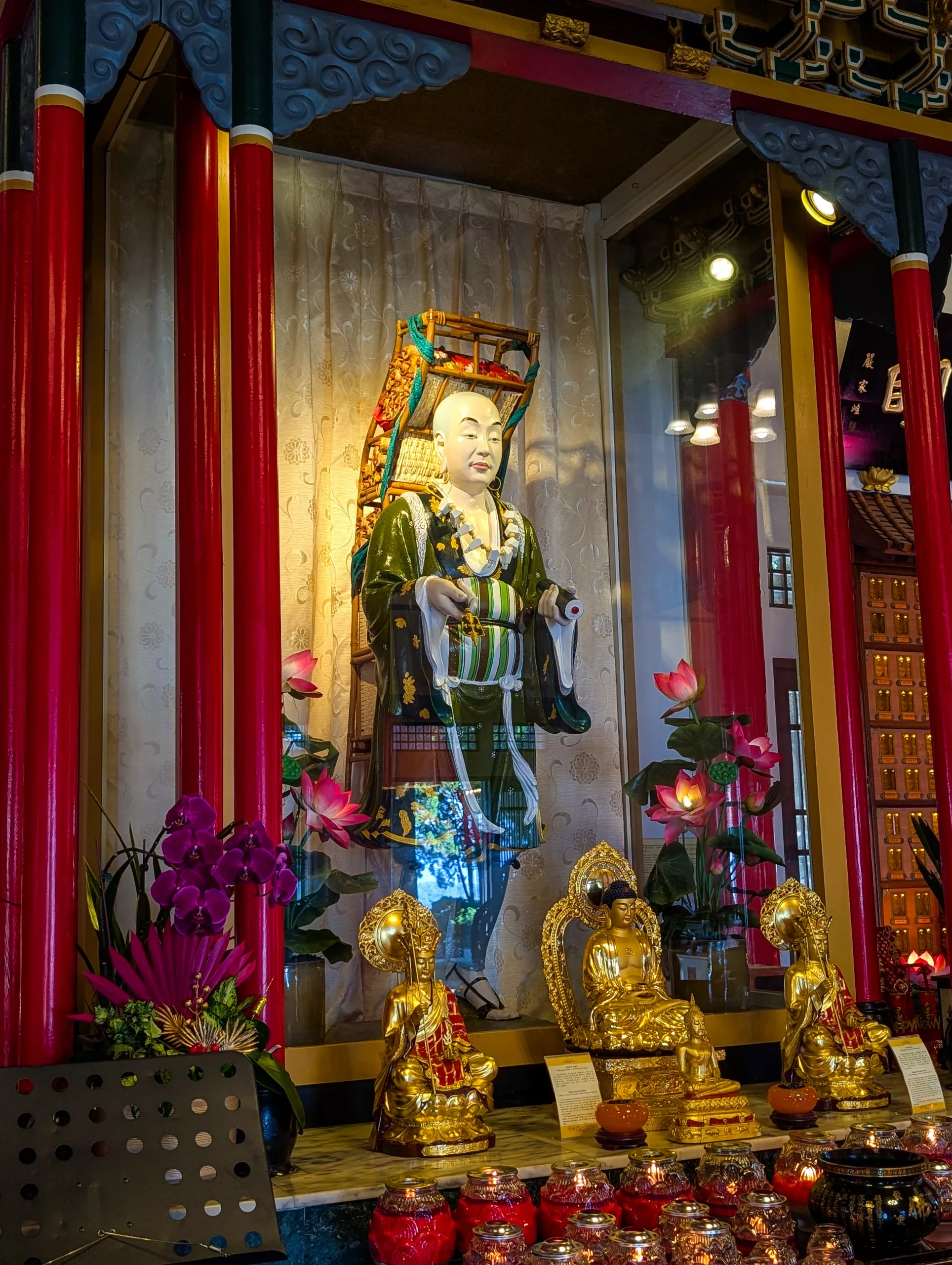 Une statue blanche vêtue de robes colorées se tient derrière une vitre, entourée de piliers rouges, de figures de Bouddha dorées, de fleurs de lotus roses et d'offrandes décoratives.