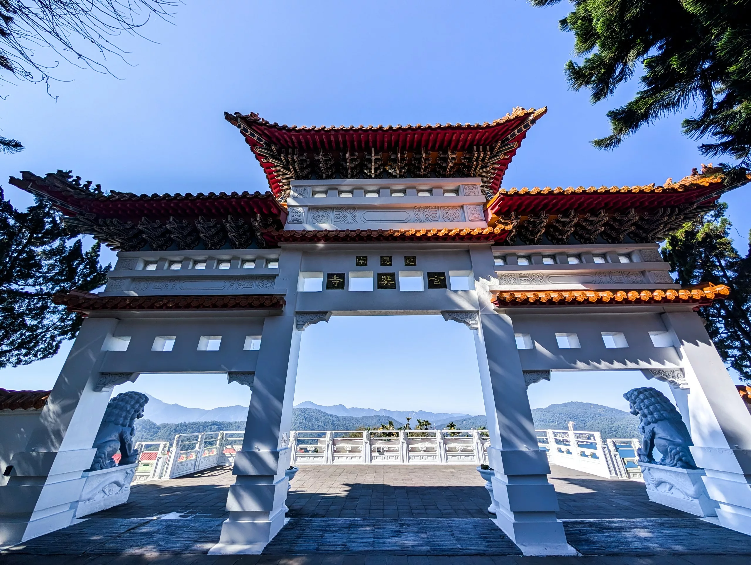 Une porte traditionnelle de style chinois avec des tuiles rouges et des piliers blancs, flanqué de lions en pierre et entouré de montagnes et d'arbres sous un ciel bleu limpide.