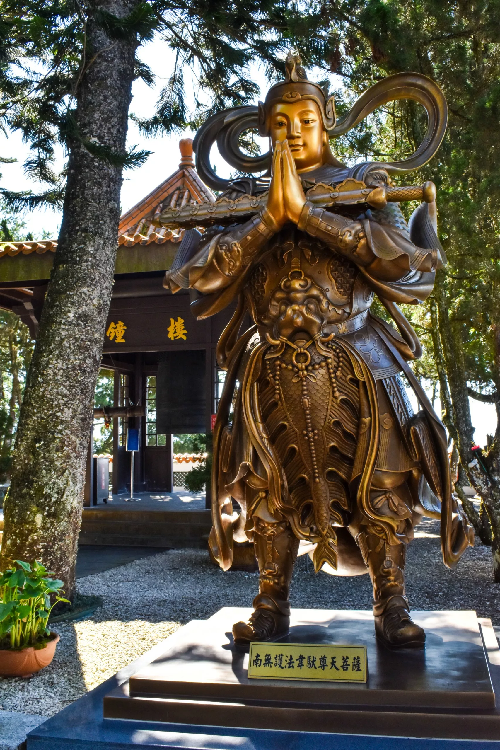 La statue en bronze d'un guerrier, les mains jointes en prière sur un piédestal au milieu des arbres, accueille les visiteurs du lac Sun Moon dans un bâtiment traditionnel avec un toit en tuiles et des signes chinois.