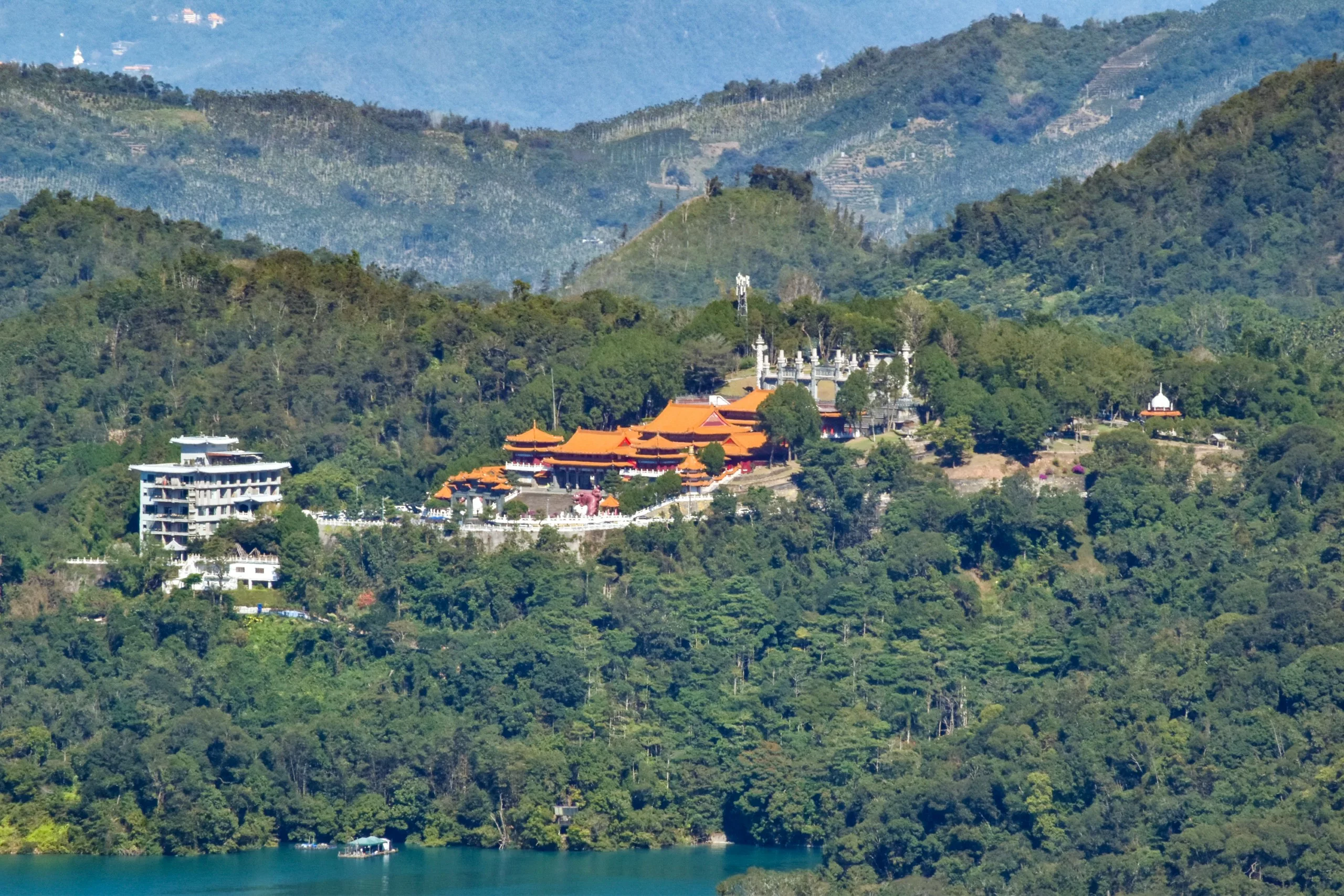 Un temple situé au sommet d'une colline, avec des toits en tuiles orange et des murs blancs, se trouve au milieu d'arbres verts denses, surplombant les montagnes
