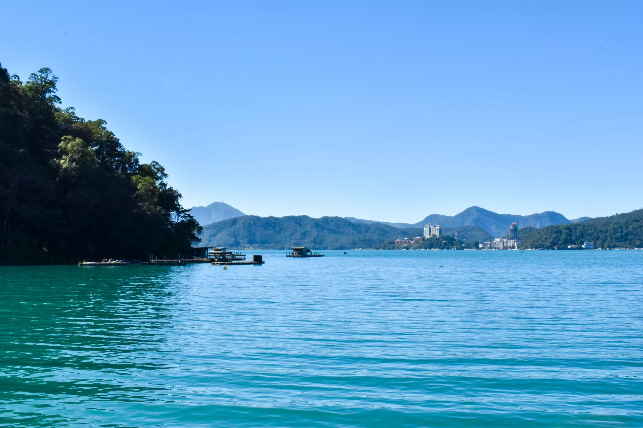 Un lac turquoise calme avec des bateaux près de la rive, entouré de collines et de montagnes couvertes d'arbres sous un ciel bleu clair. Des bâtiments sont visibles au loin.