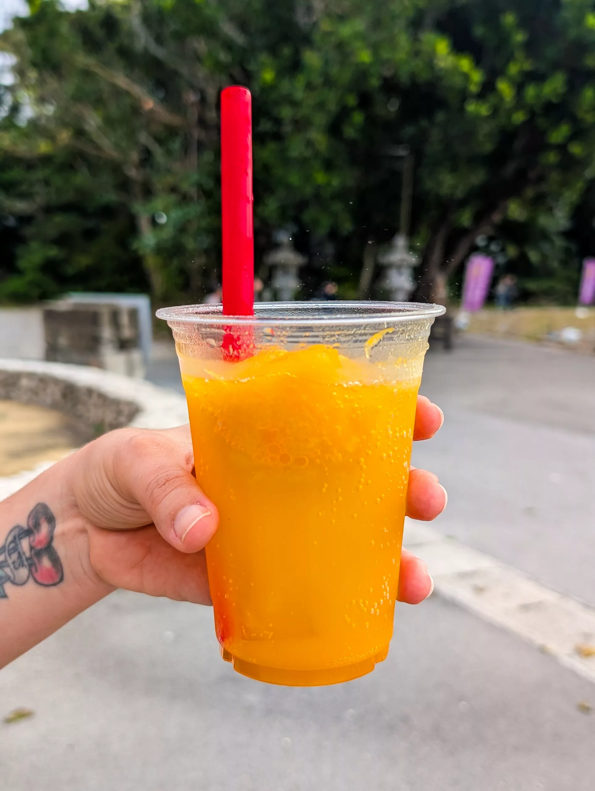 Une main tenant un gobelet en plastique transparent rempli d'une boisson gazeuse couleur orange et de glaçons, avec une grande paille rouge