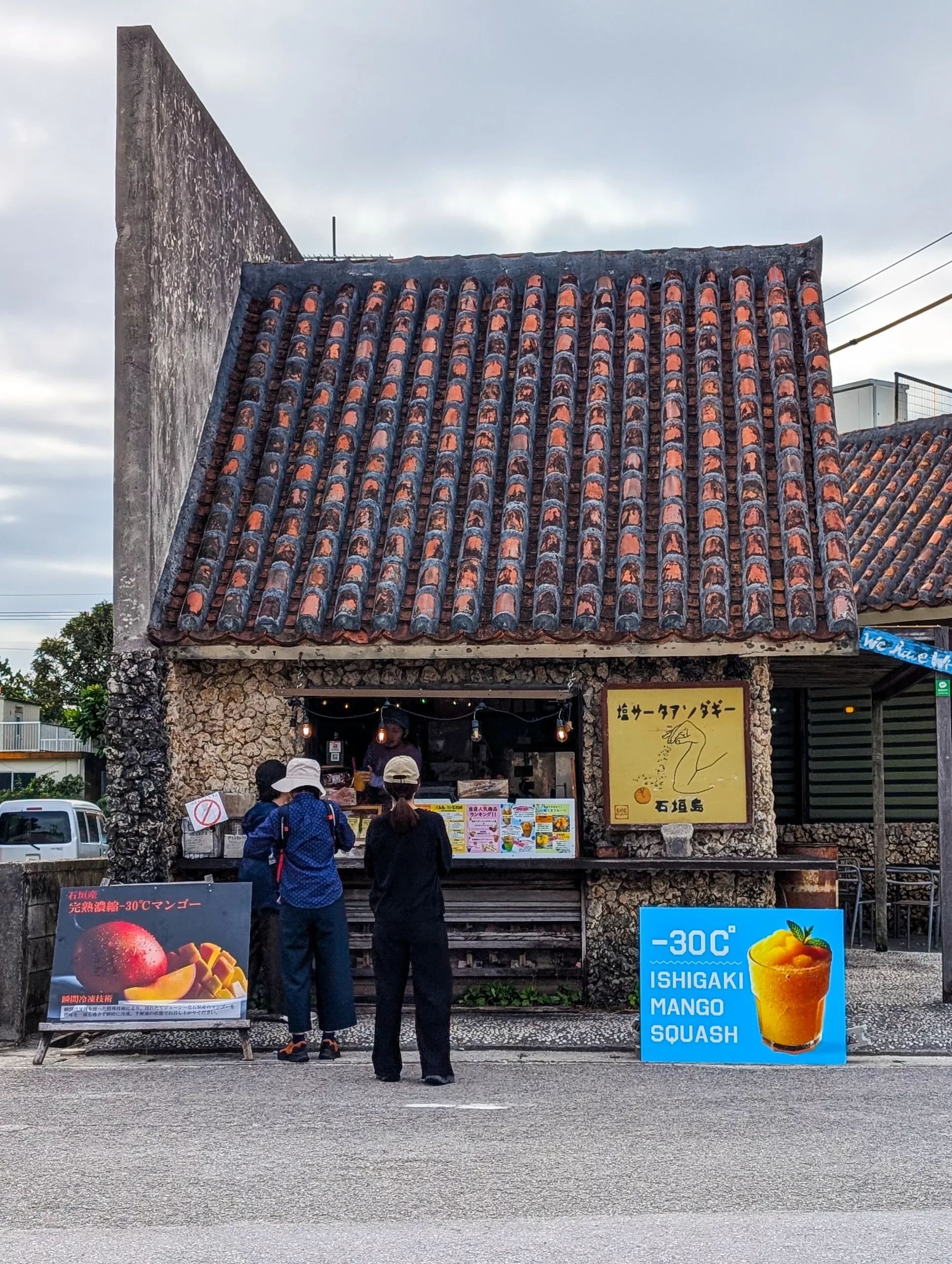 Trois personnes se tiennent près d'un petit bâtiment en pierre au toit de tuiles rouges. Des panneaux annoncent de la purée de mangue d'Ishigaki servie à -30 °C