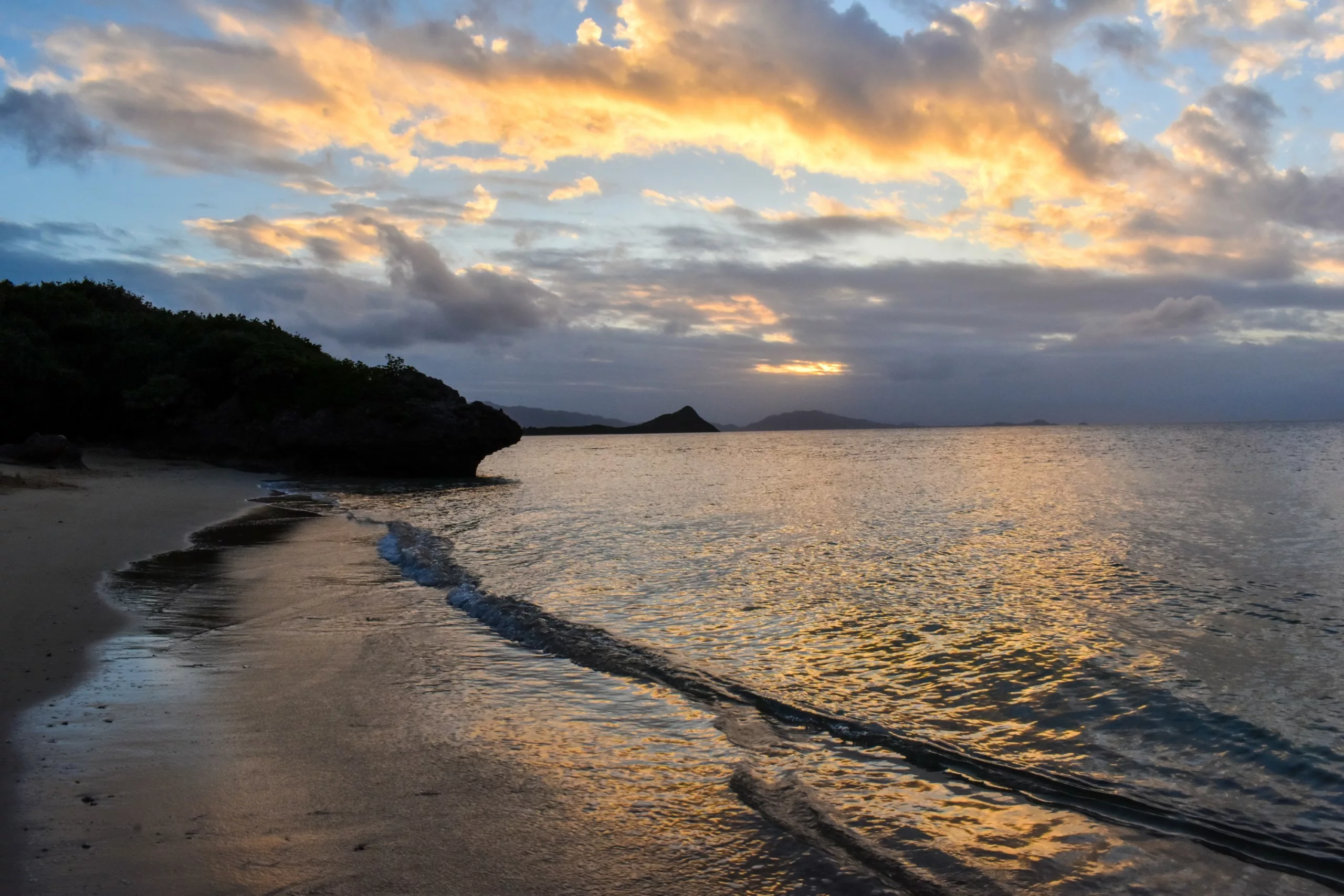 Une plage de sable aux vagues douces au coucher du soleil, les nuages se teintent d'orange sur l'eau calme, tandis que les collines lointaines se fondent dans un affleurement rocheux sur la gauche.