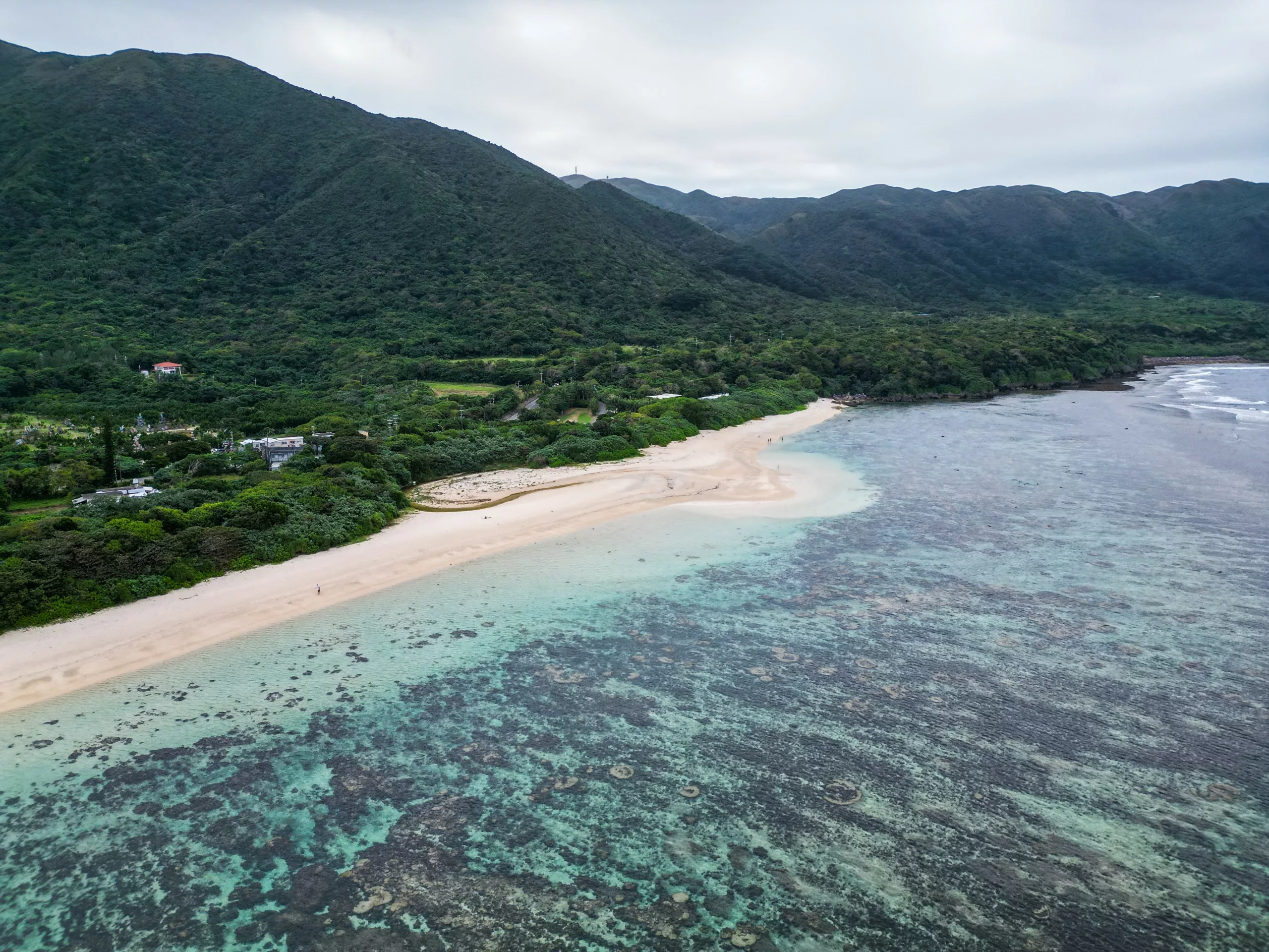 Vue aérienne d'une plage de sable aux eaux turquoise cristallines à Ishigaki, entourée d'une végétation verdoyante et de collines boisées sous un ciel nuageux