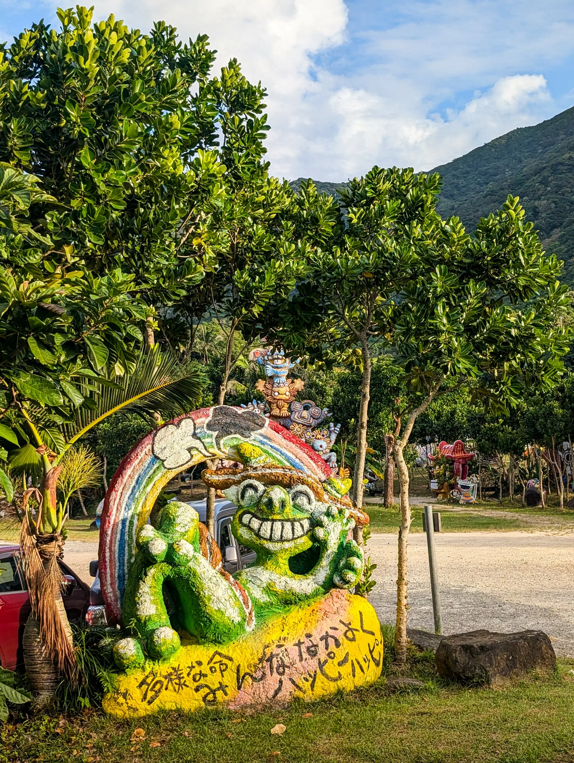 Une statue colorée verte au large sourire, adossée à un arc-en-ciel, accueille les visiteurs à côté d’un parking en gravier. Un texte en japonais orne son socle