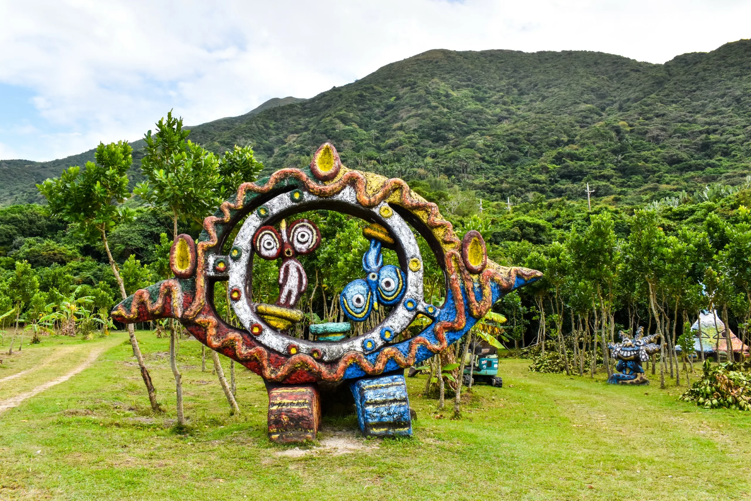 Une grande sculpture colorée ornée de visages et de motifs abstraits, se dresse sur un terrain herbeux, avec en toile de fond des arbres et des montagnes.