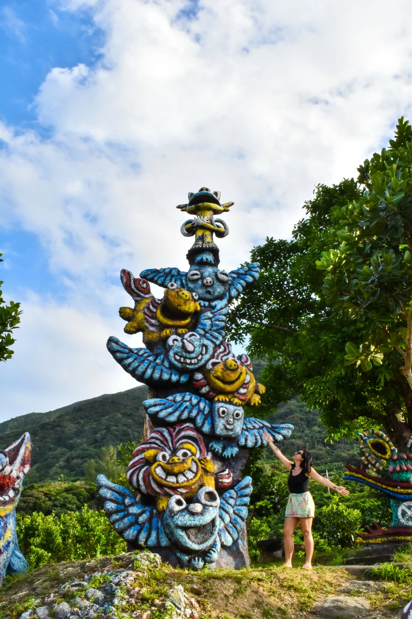 Une femme se tient à côté d’une grande sculpture colorée, semblable à un totem, composée de créatures fantaisistes empilées les unes sur les autres