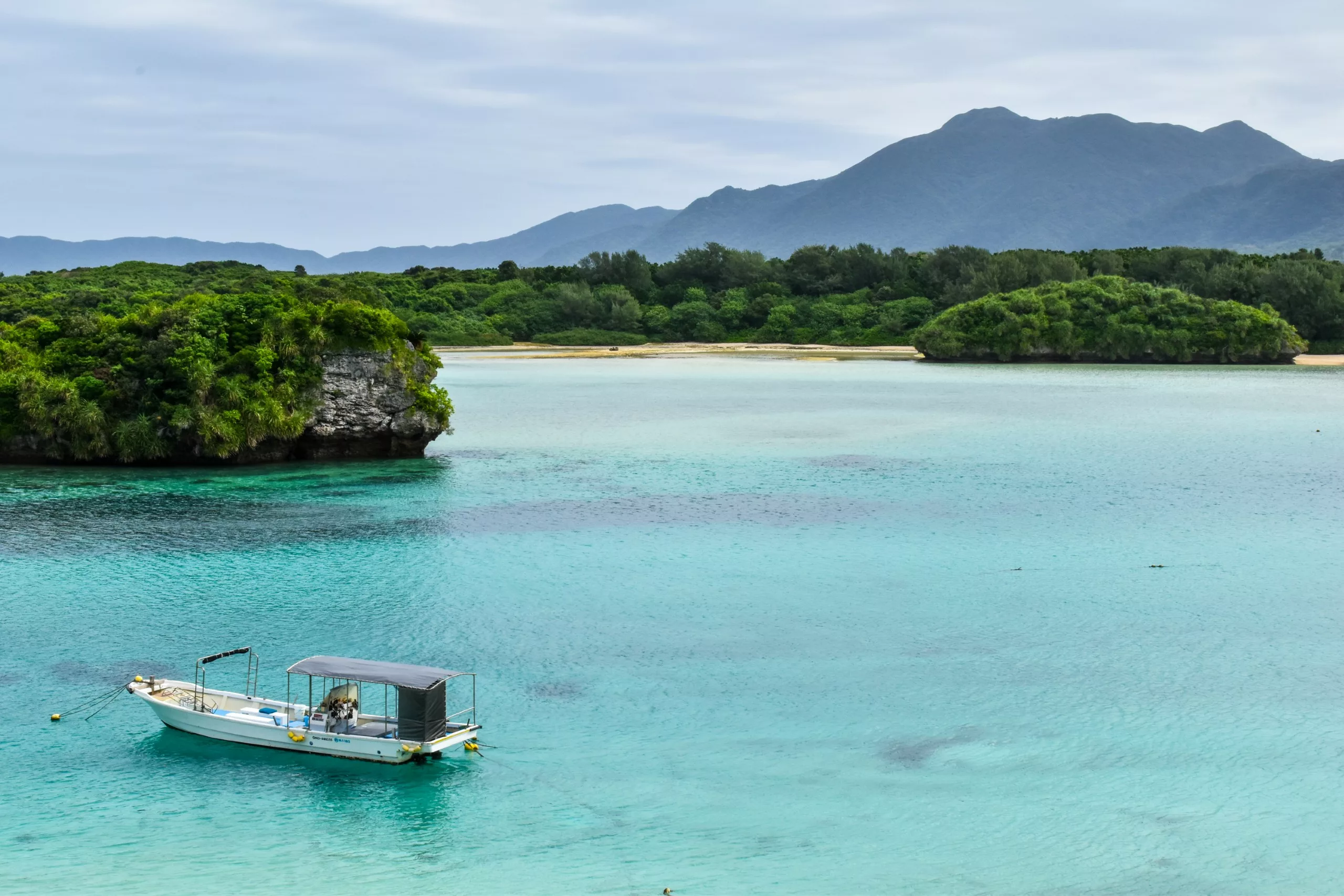 Un petit bateau blanc flotte sur une eau turquoise et limpide de la baie de Kabira à Ishigaki près d'une île verdoyante et boisée