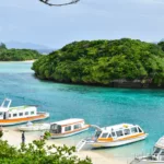 De petits bateaux blancs et orange sont amarrés le long d'une plage de sable d'Ishigaki bordée d'une eau turquoise cristalline avec en arrière-plan des îles verdoyantes sous un ciel légèrement couvert.