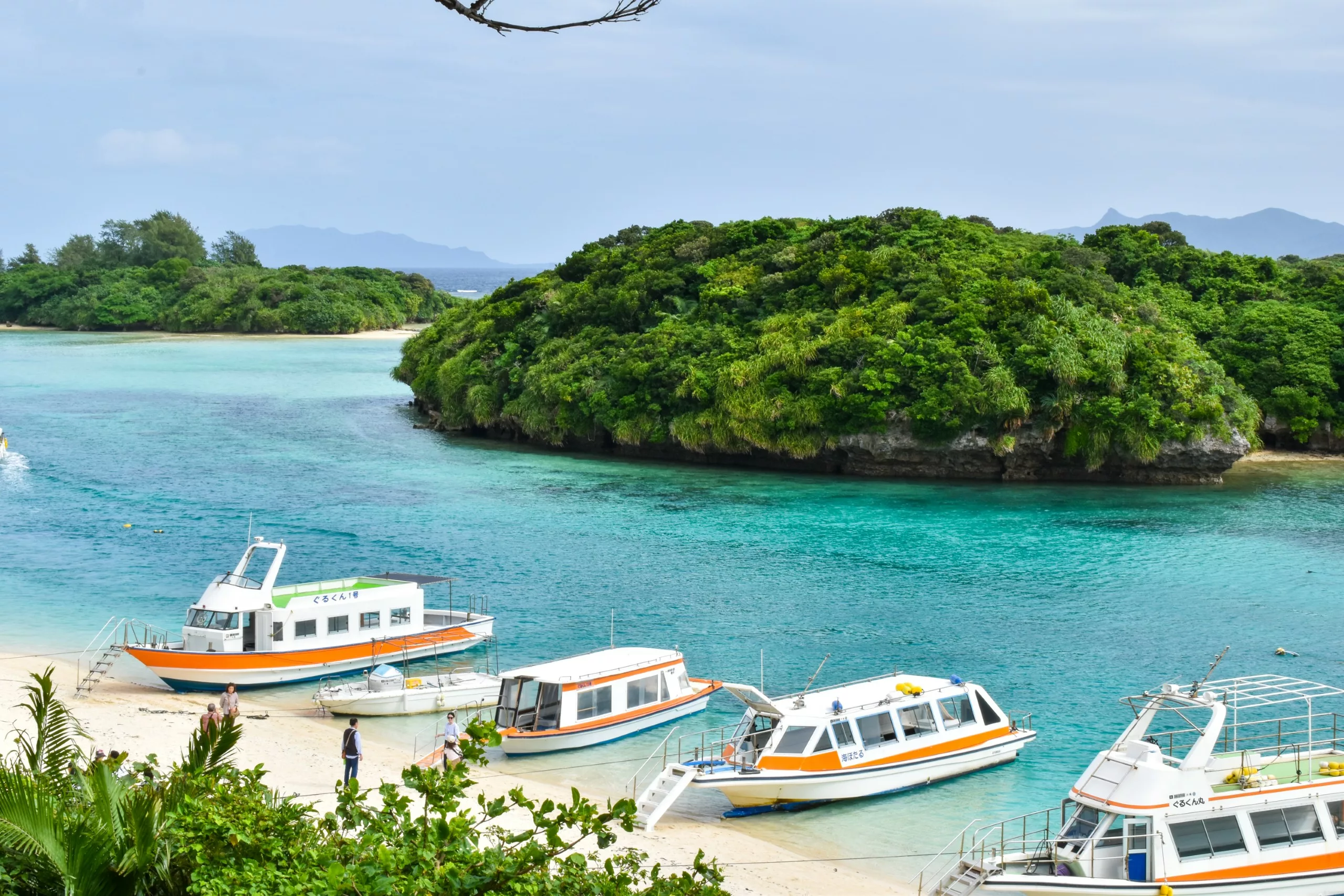 De petits bateaux blancs et orange sont amarrés le long d'une plage de sable d'Ishigaki bordée d'une eau turquoise cristalline avec en arrière-plan des îles verdoyantes sous un ciel légèrement couvert.