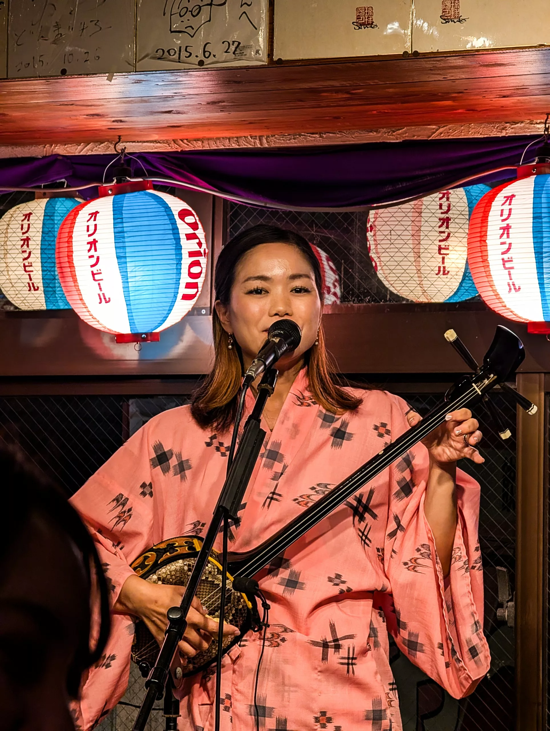 Une femme vêtue d'un kimono rose joue d'un instrument à cordes traditionnel et chante dans un micro. Derrière elle, des lanternes rouges, blanches et bleues brillent.