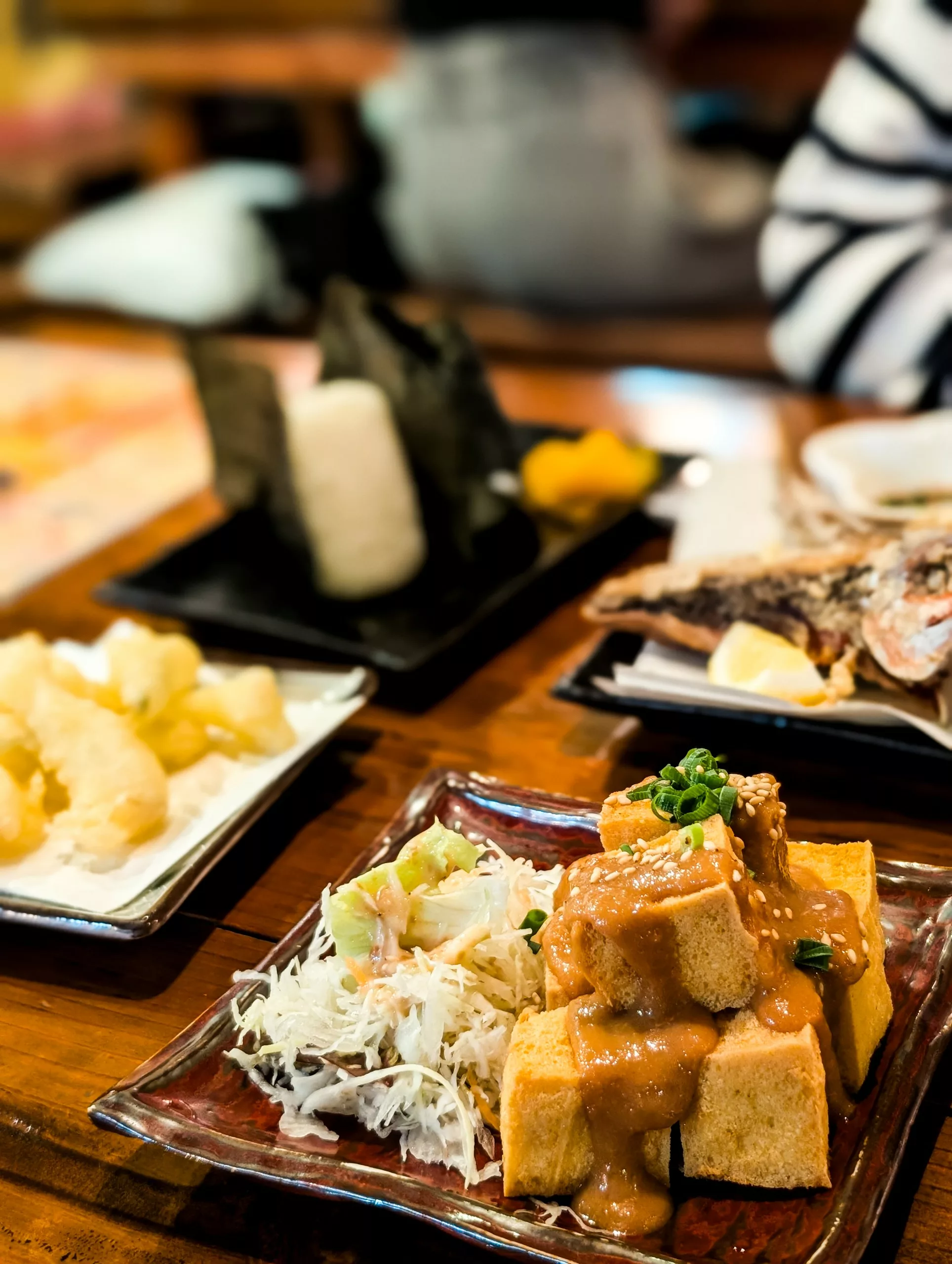 Une table en bois garnie d'assiettes de plats japonais — tels que du tofu frit, du tempura, une boulette de riz et du poisson grillé.