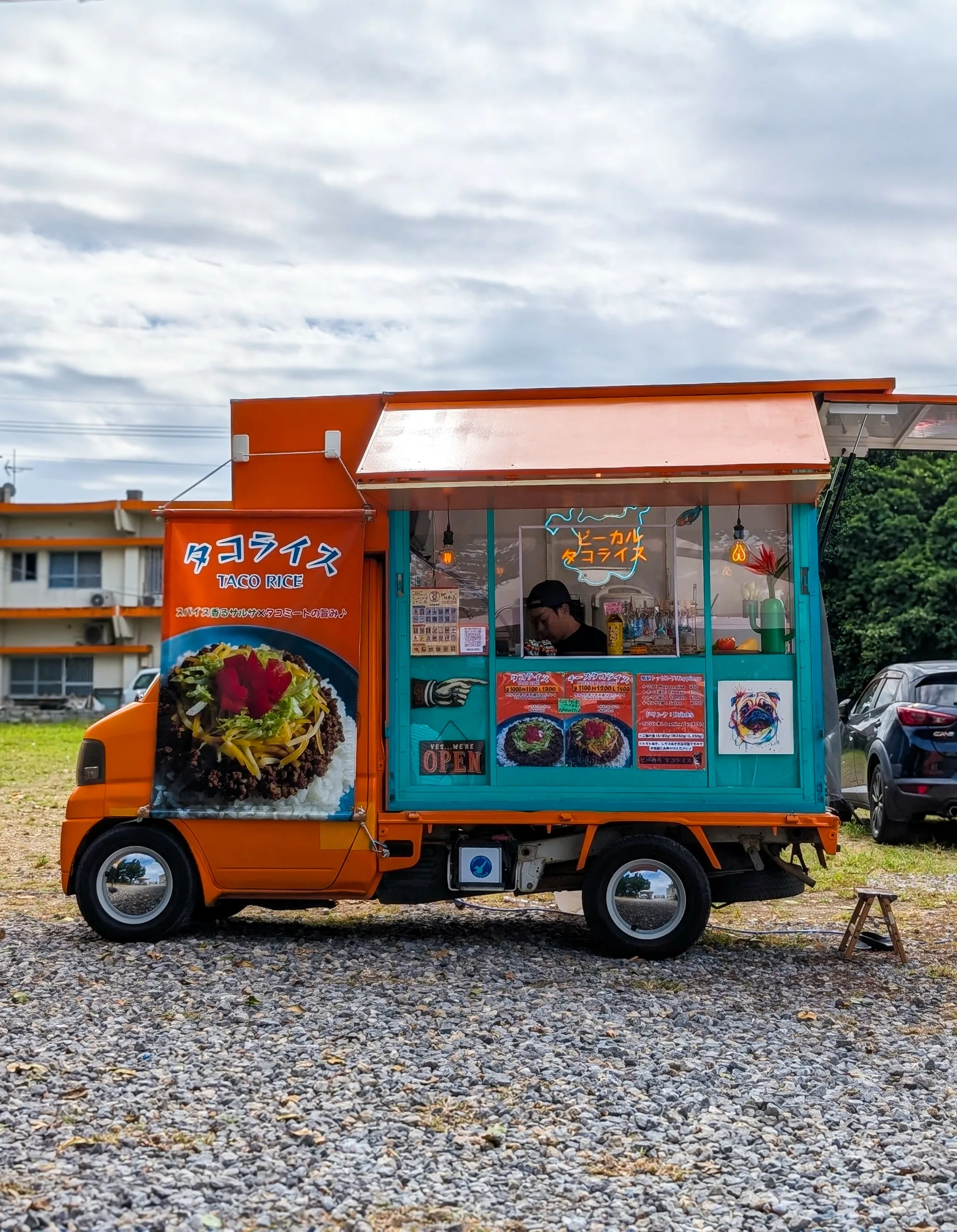 Un petit food truck orange et turquoise, orné d'enseignes indiquant les plats proposés, est garé sur du gravier, prêt à servir.