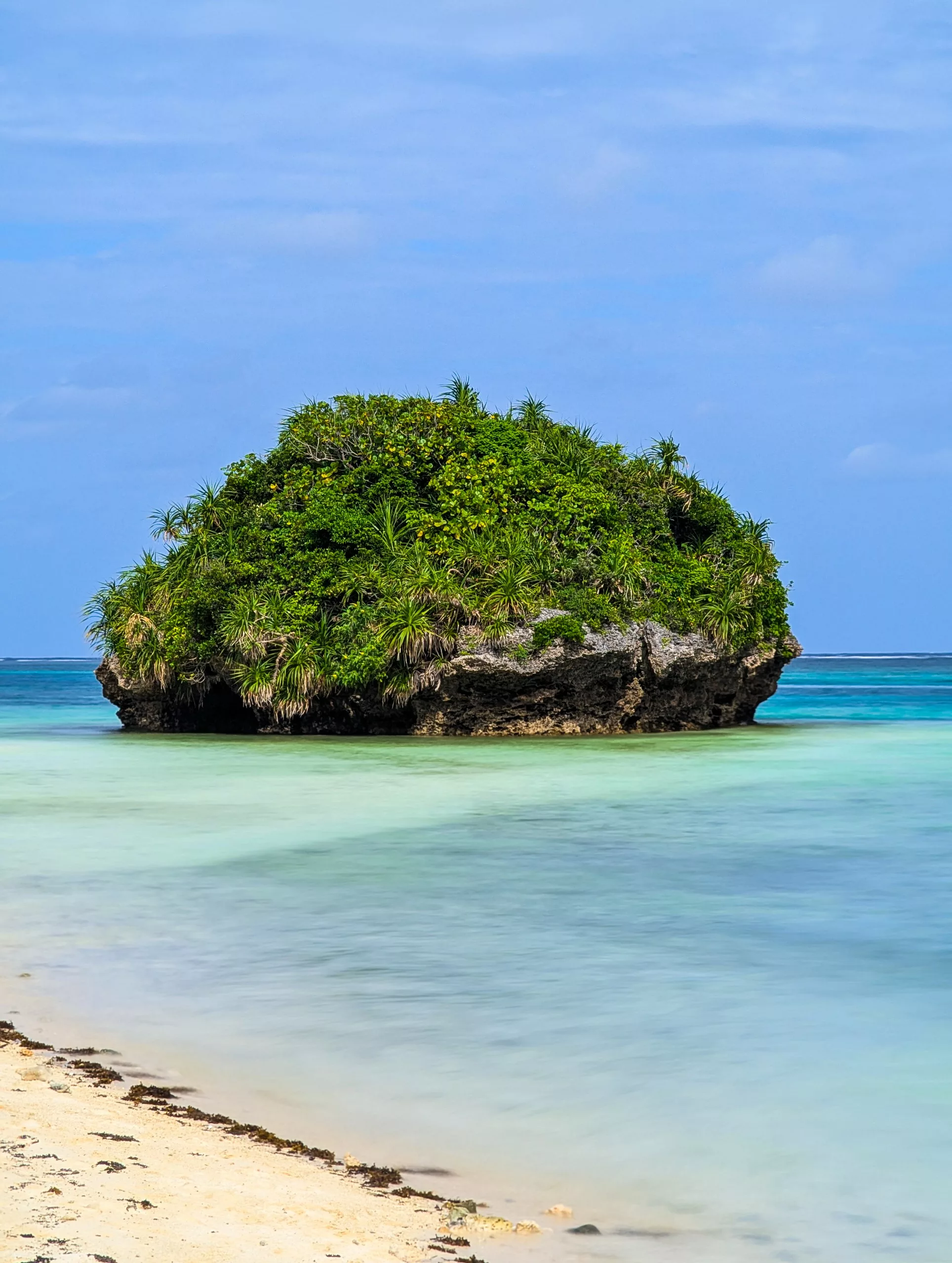 Une petite île rocheuse recouverte d'une végétation dense et verdoyante se dresse dans des eaux turquoise claires et peu profondes, près d'une plage de sable, sous un ciel d'un bleu éclatant