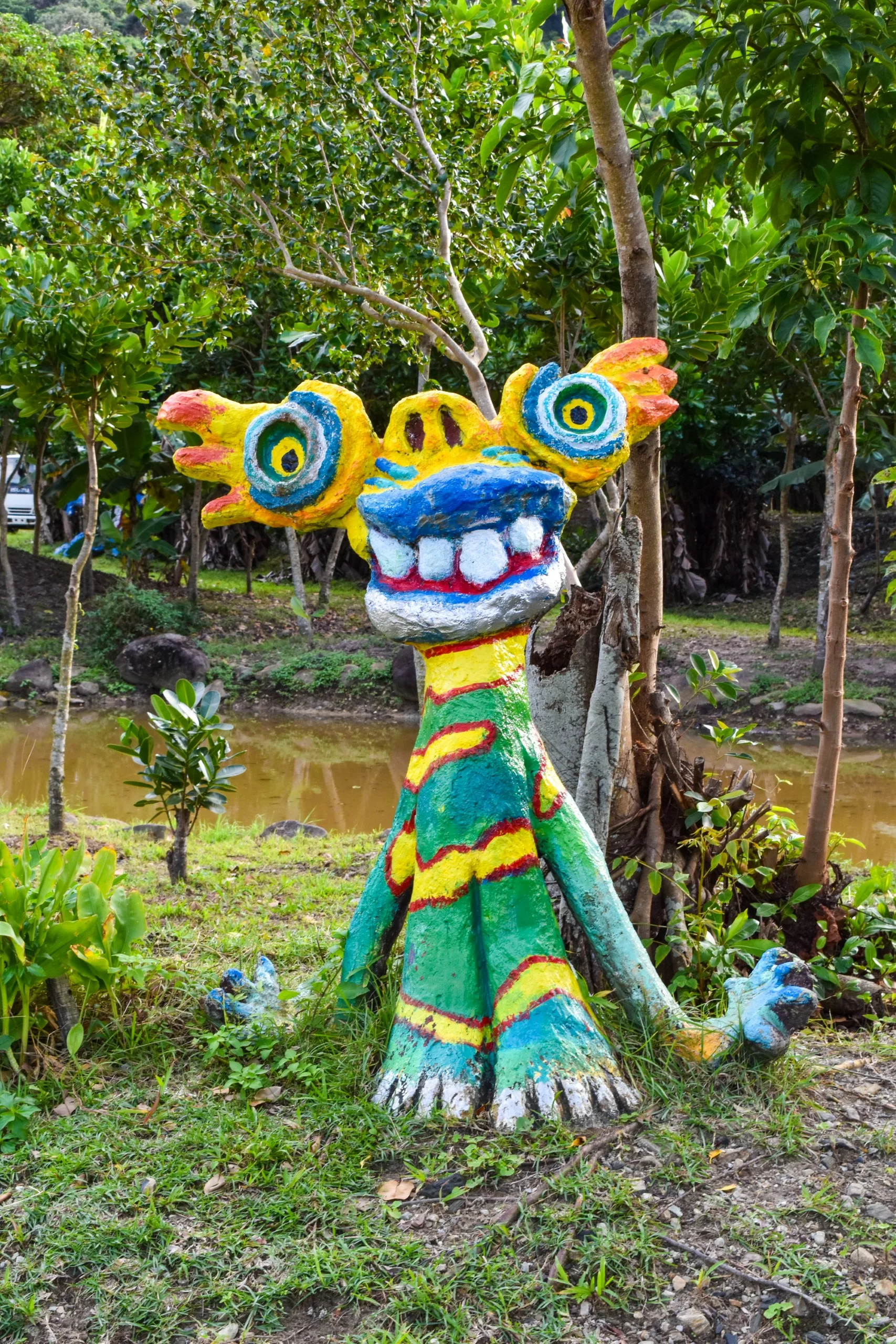 Une sculpture colorée et fantaisiste représentant une créature aux grands yeux, aux grandes dents et aux motifs rayés se dresse sur l'herbe, près des arbres et d'un étang brun