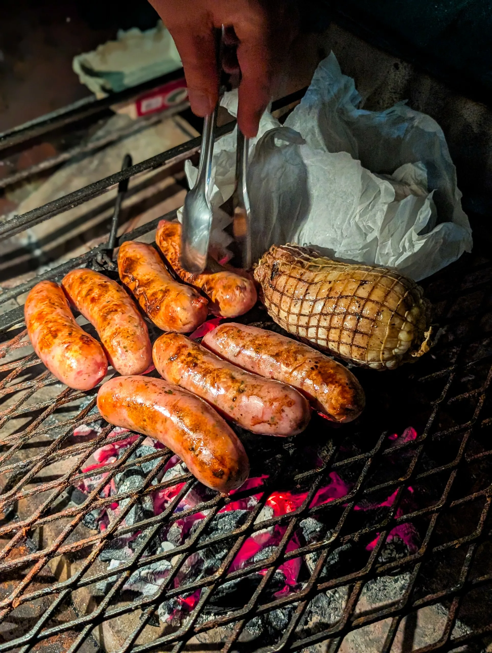 Un barbecue avec des grosses saucisses qui grillent et un espèce de roti. Une spatule touchent les saucisses pour les retourner.