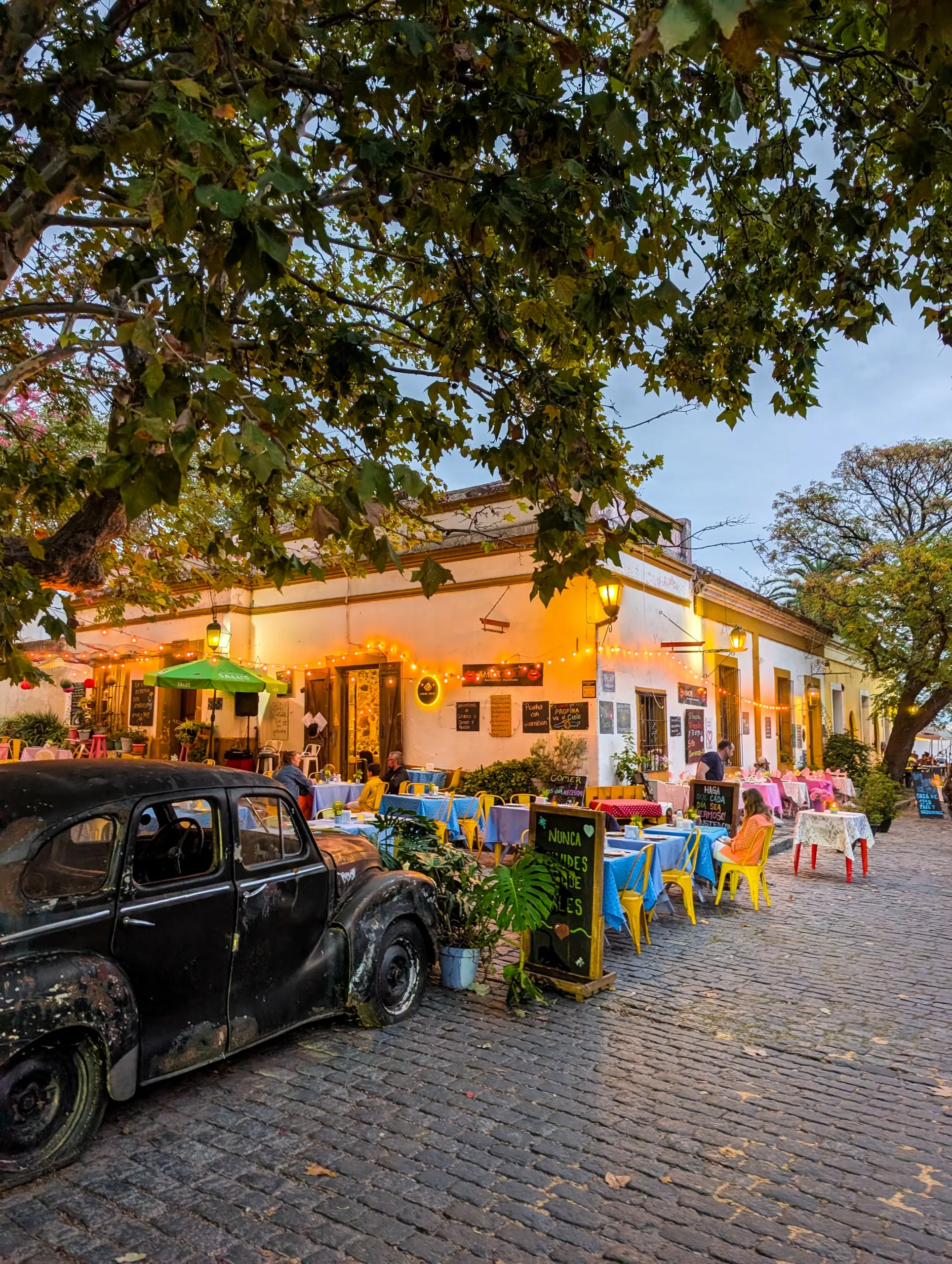 Une rue pavée de Colonia del Sacramento, où une vieille voiture noire est garée à côté d'un café. Des tables en terrasse, entourées de chaises colorées, sont disposées à l'ombre de grands arbres et baignées d'une lumière chaleureuse, tandis que les clients dînent au crépuscule près d'un menu écrit à la craie sur un tableau noir.