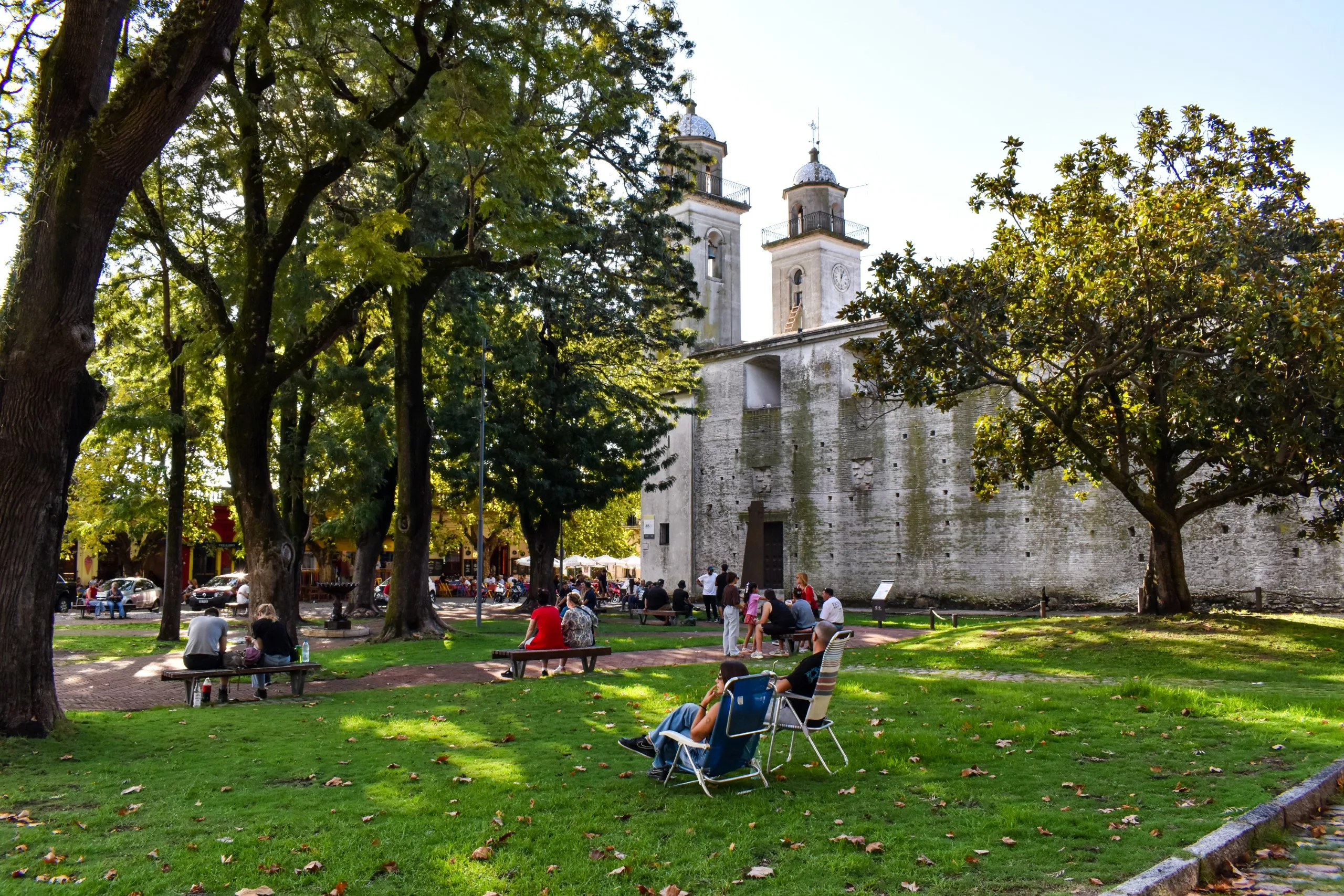 Dans un parc ombragé situé près de d'une église historique en pierre dotée de deux clochers, des gens sont assis sur des bancs et des chaises de jardin. De grands arbres procurent de l'ombre et les feuilles mortes recouvrent l'herbe.