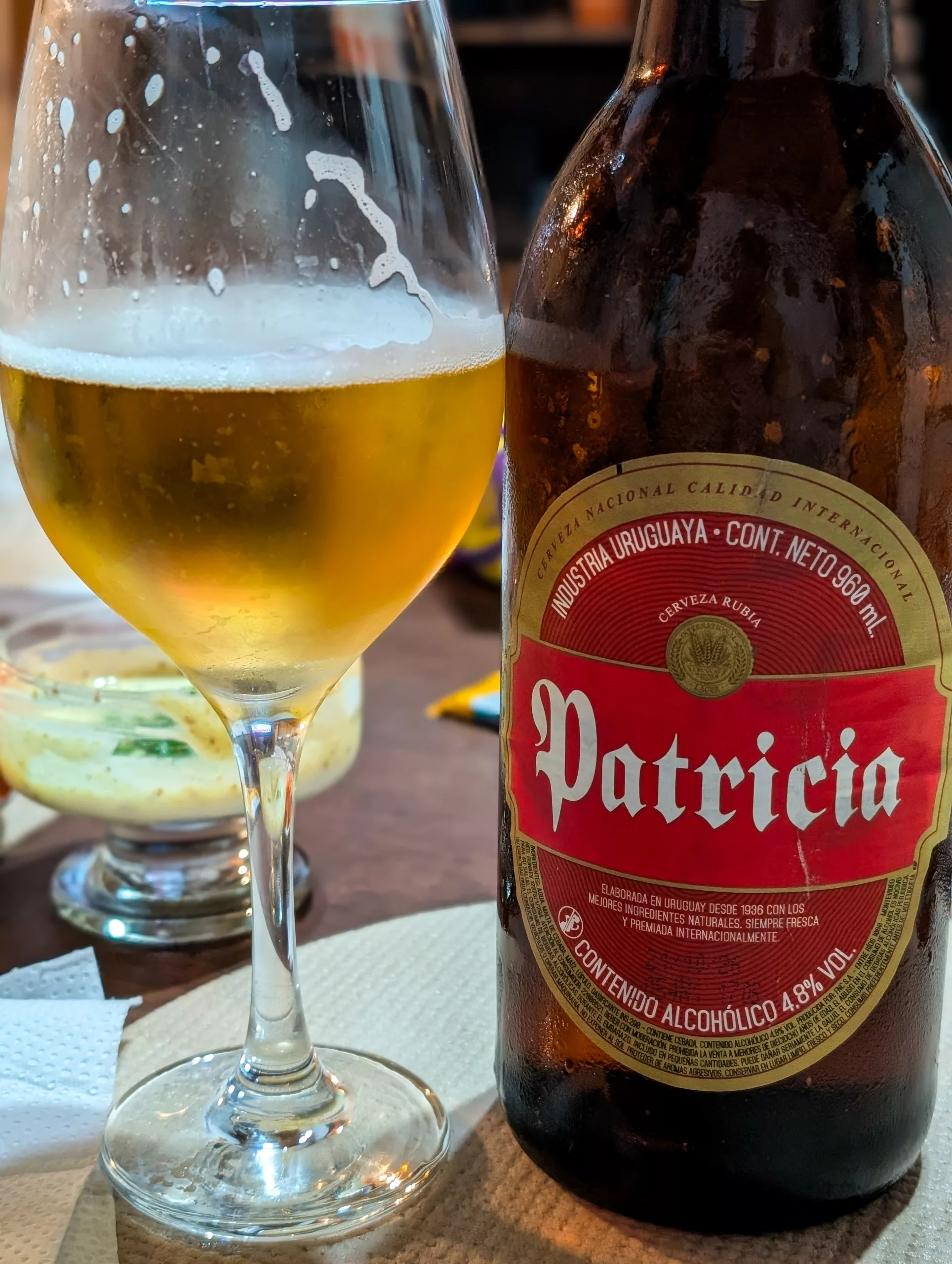 Un verre de bière avec des traces de mousse est posé à côté d'une bouteille brune portant l'étiquette « Patricia » sur une table avec en arrière plan une serviette beige et un petit bol de sauce.