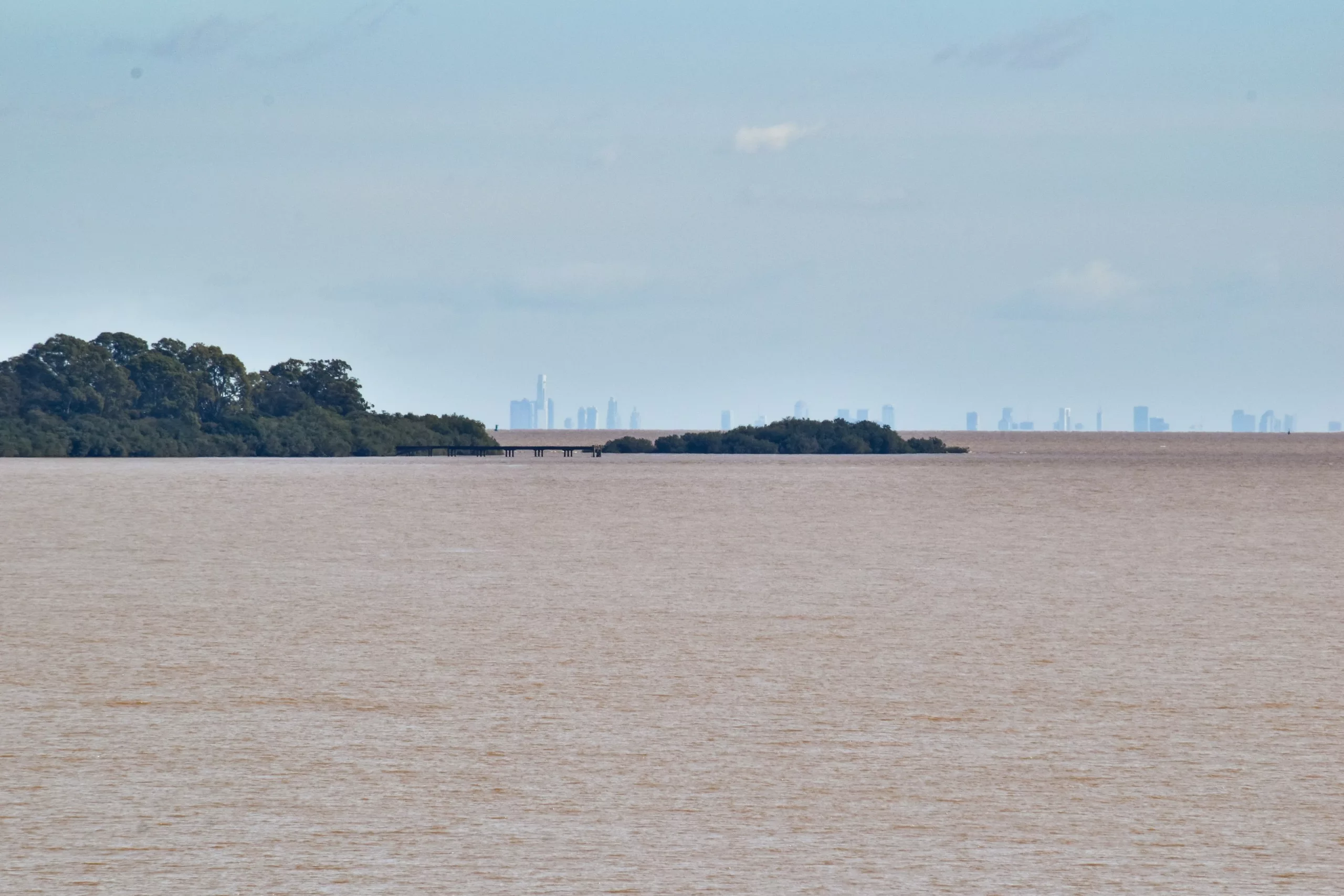 Un large fleuve aux eaux brunâtres près de Colonia del Sacramento, de petites îles verdoyantes et la silhouette de la ville de Buenos Aires, visible au loin à l'horizon.