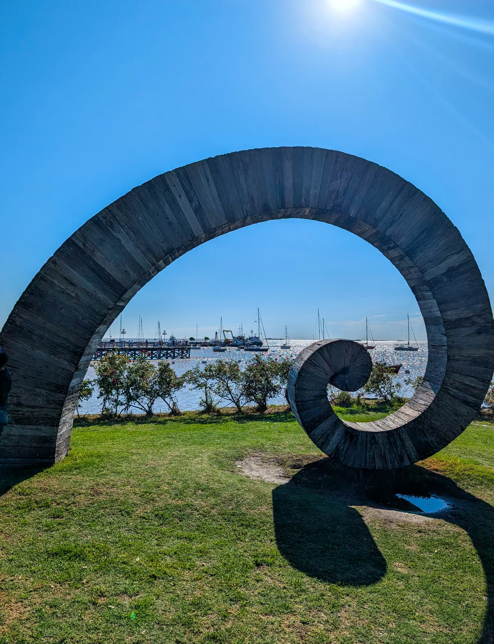 Une grande sculpture en spirale trône sur la pelouse près de l'eau, projetant son ombre. Des voiliers sont amarrés à l'arrière-plan, et le ciel est dégagé, sous un soleil radieux.