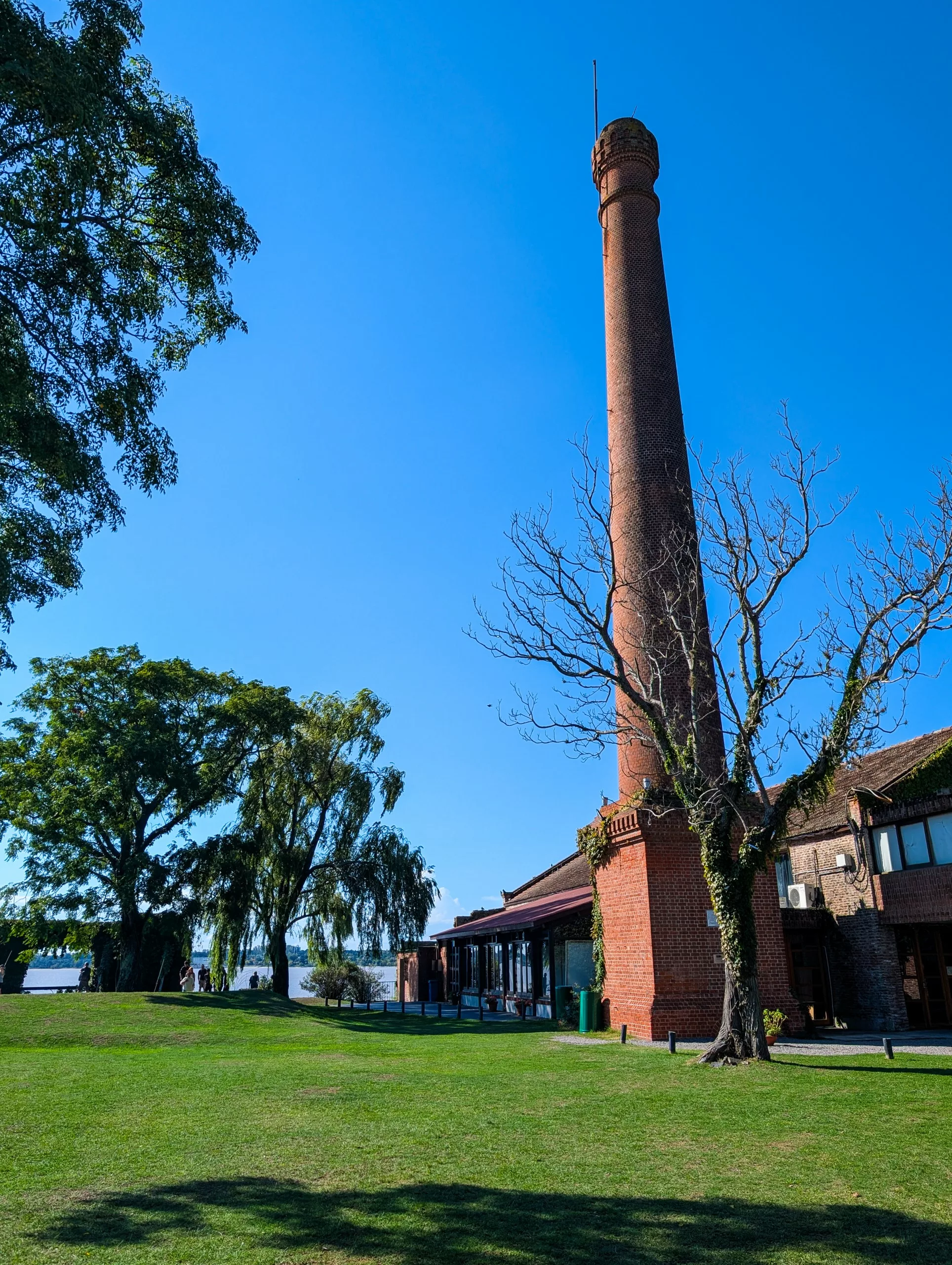 À Colonia del Sacramento, une haute cheminée en briques se dresse à côté d'un bâtiment aux grandes fenêtres, au milieu d'une pelouse verte, d'arbres et sous un ciel bleu.
