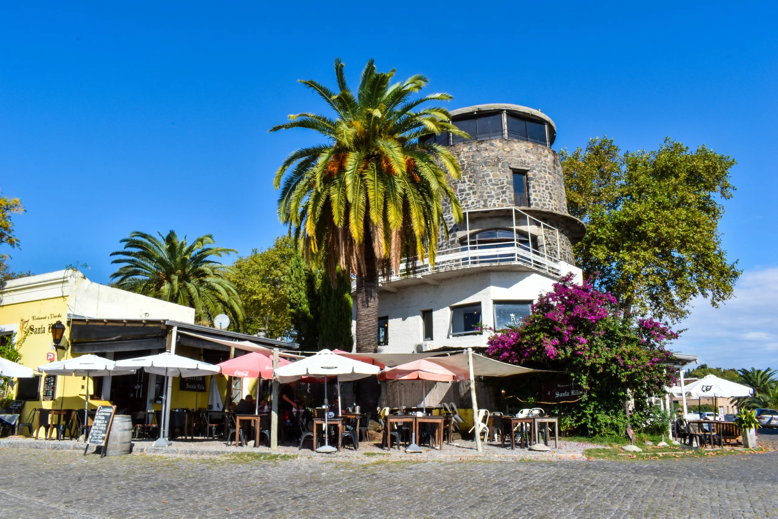 Terrasse de café avec des tables et des parasols dans une rue pavée de Colonia del Sacramento, à côté d'un bâtiment en pierre ressemblant à une tour, entourée de palmiers et d'arbustes à fleurs violettes sous un ciel bleu limpide.